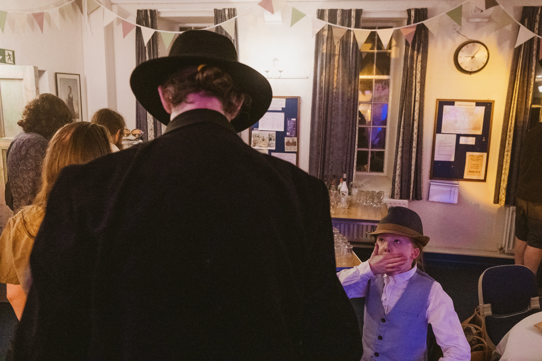 Child in hat covers mouth at indoor event.