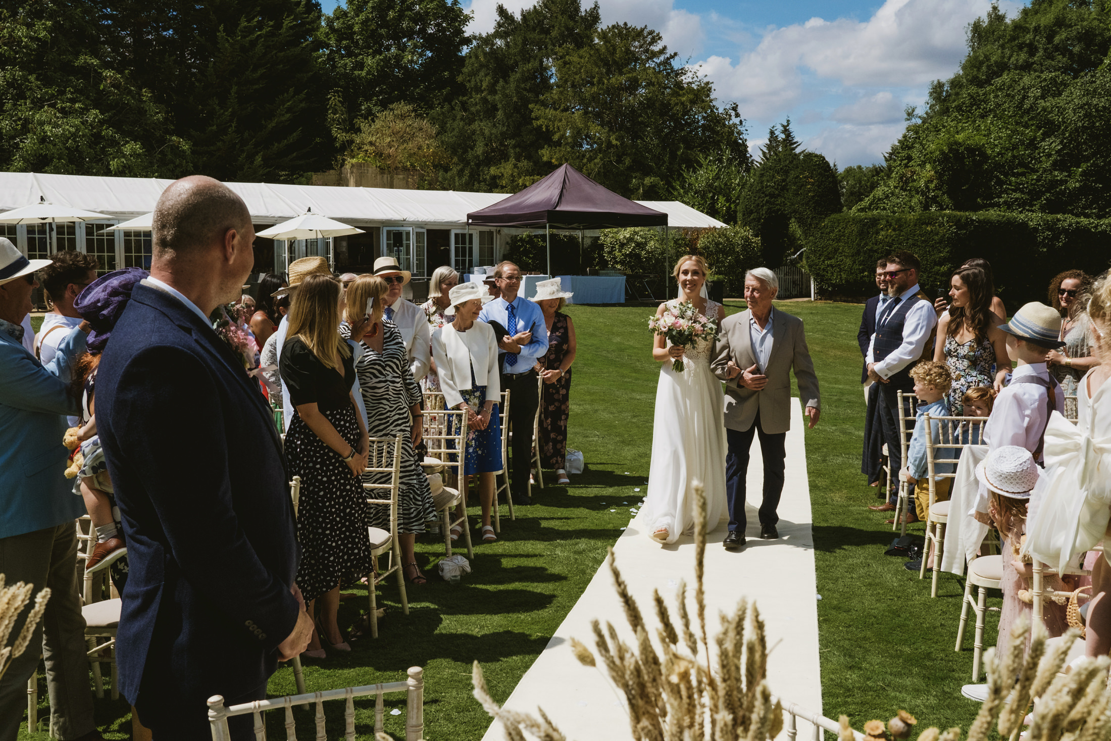 Bride walking down outdoor aisle with guests seated.
