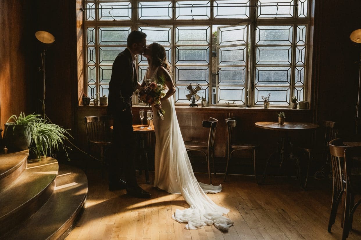 A graphic silhouette of the bride and groom kissing in a light-drenched window at the Town Hall Hotel. This documentary wedding photograph utilizes the venue's high-contrast, directional light to create a cinematic, artistic frame during the reception.