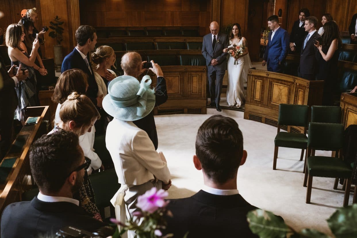 A documentary photograph of the bride’s entrance into the Council Chamber at the Town Hall Hotel. The shot captures the quiet anticipation and the dramatic scale of the circular ceremony room as the processional begins.