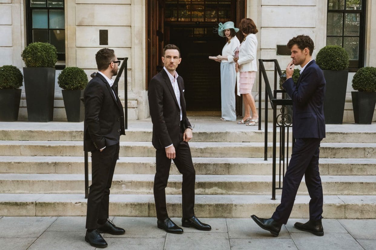A candid documentary photograph of the groom and wedding guests sharing a laugh on the front steps of the Town Hall Hotel. Captured against the backdrop of Patriot Square, this shot highlights the real, unscripted human interactions and the iconic exterior of the Bethnal Green venue.
