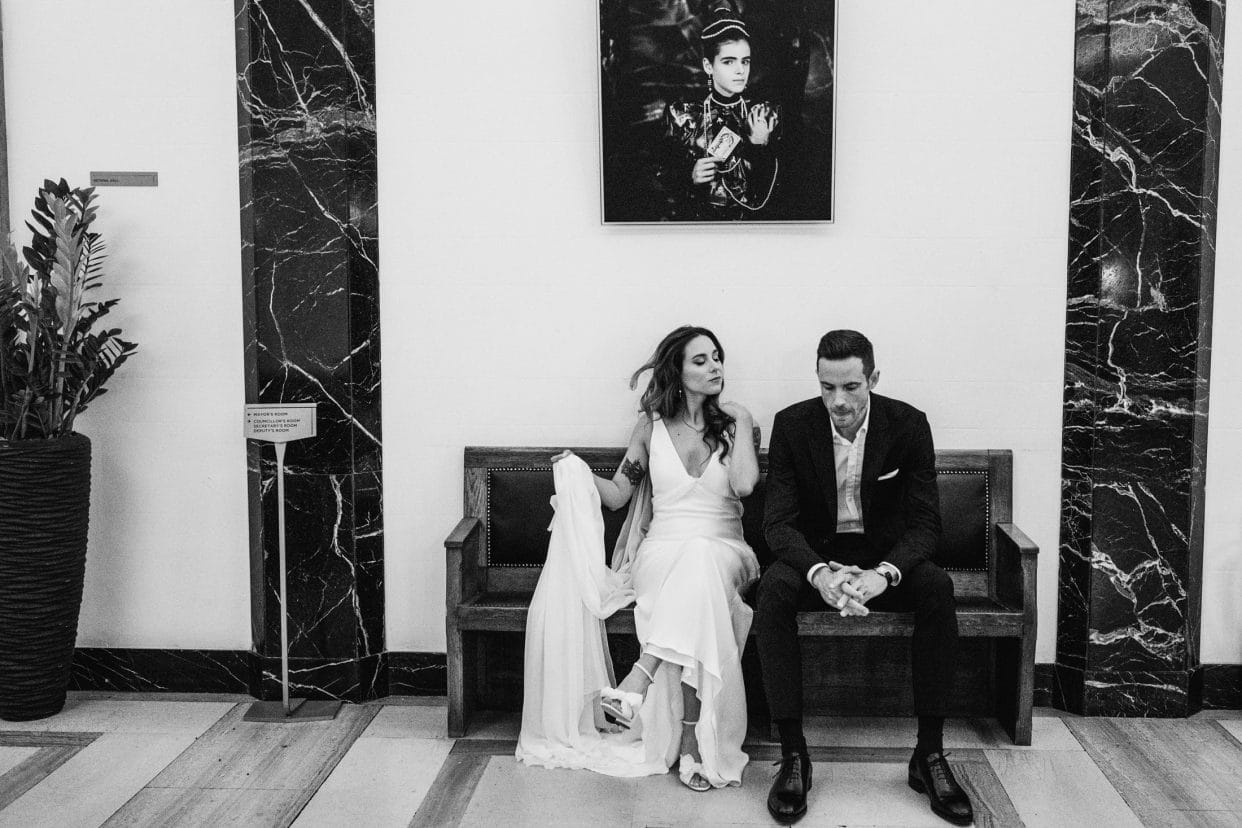 A layered documentary photograph of the bride and groom sitting inside the Town Hall Hotel, framed by a large overhead painting. This unposed moment captures the couple within the venue’s artistic, avant-garde interior, treating the hotel's heritage as a silent character in the story.