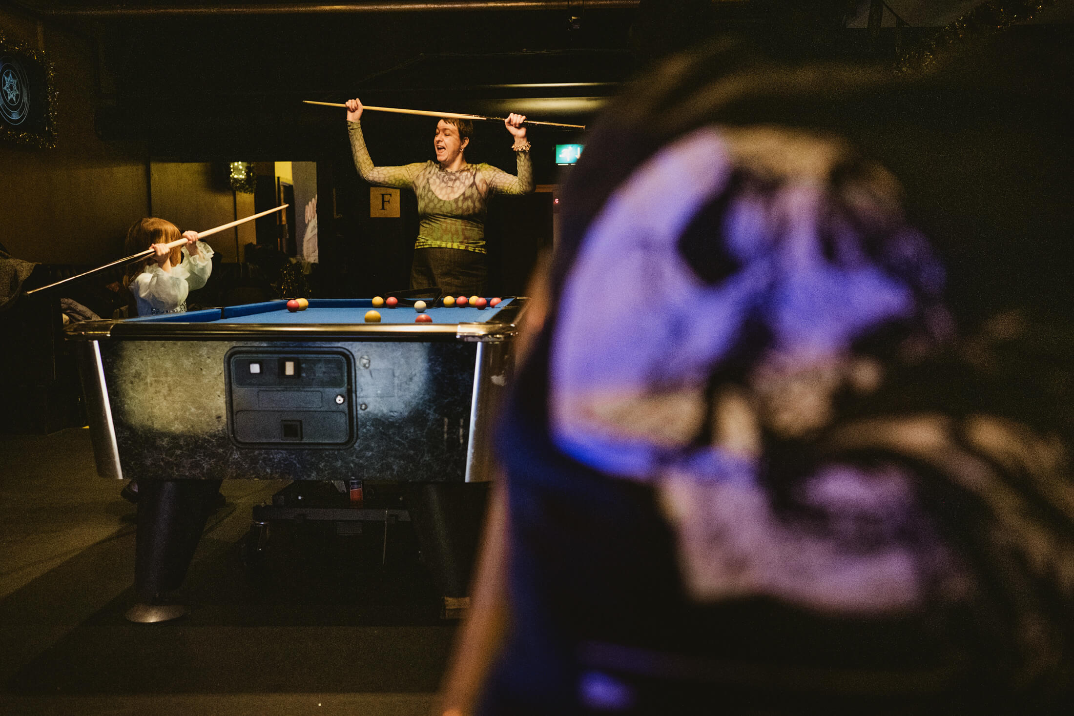Guests play pool at a lively wedding reception, one cheering with cue raised above the table.