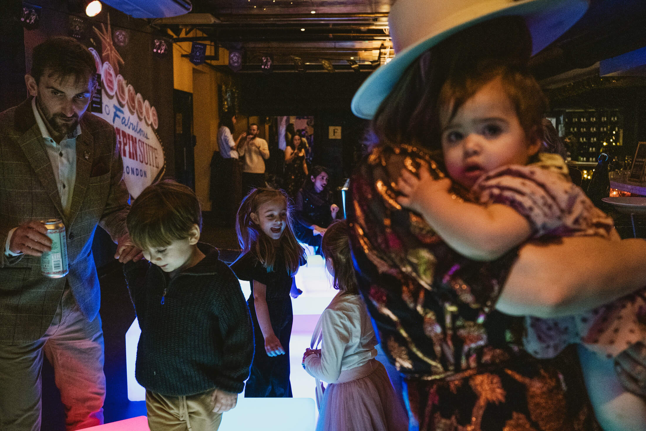 Children dance on glowing floor as woman cradles toddler during wedding reception celebrations.