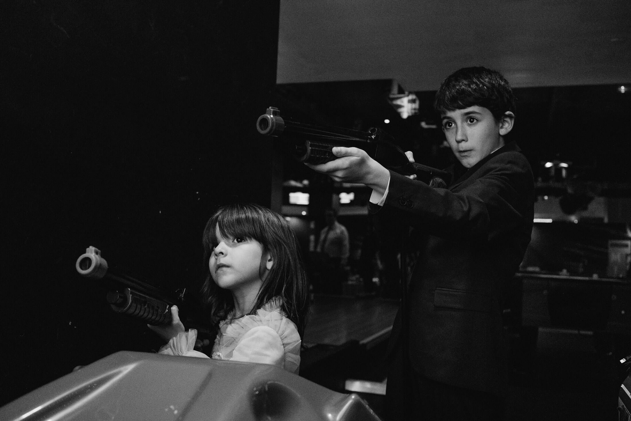 Two children in formal wear play arcade shooting game at wedding reception, eyes wide with concentration.