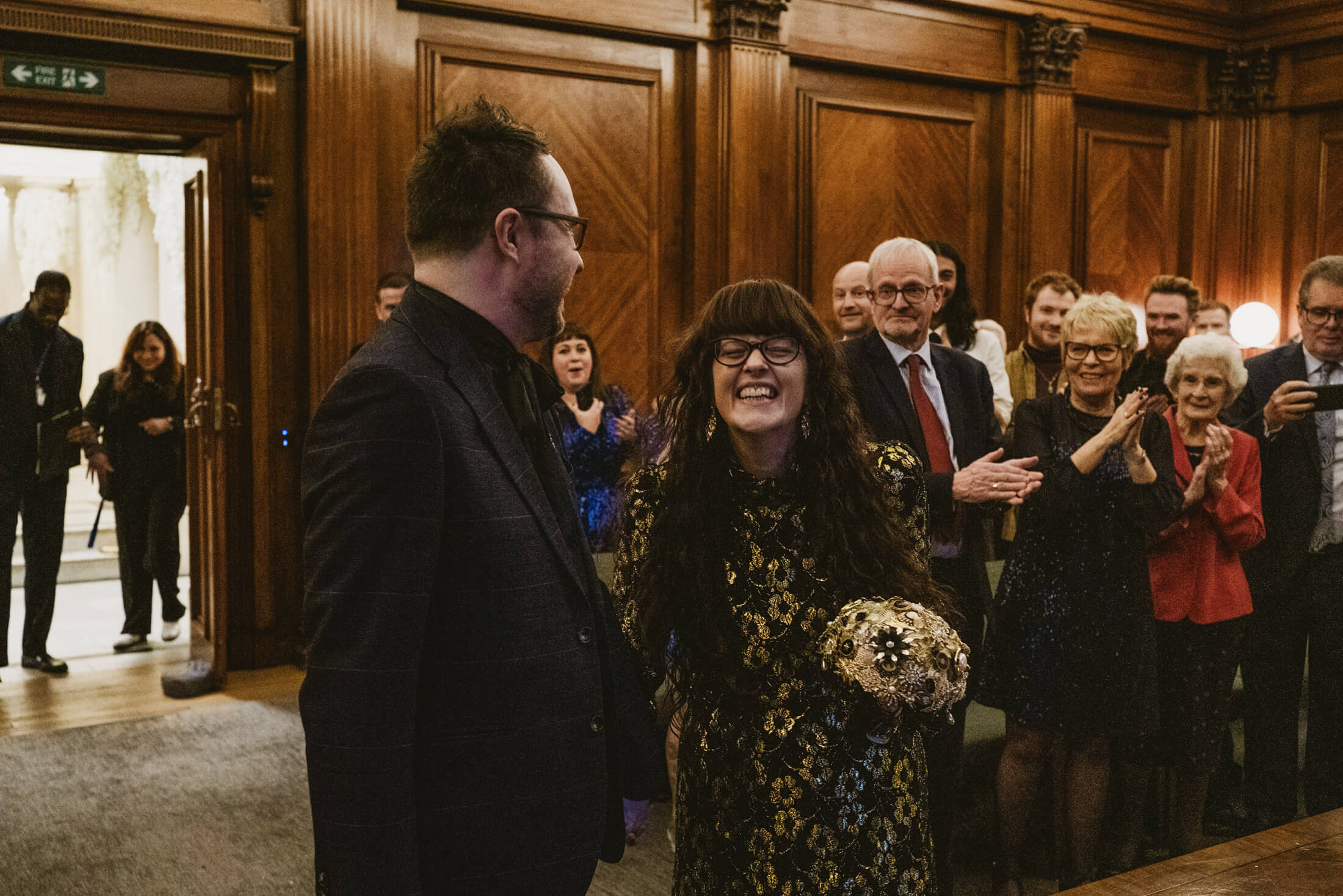 Bride laughs holding bouquet as guests applaud in wood-panelled room during wedding celebration.