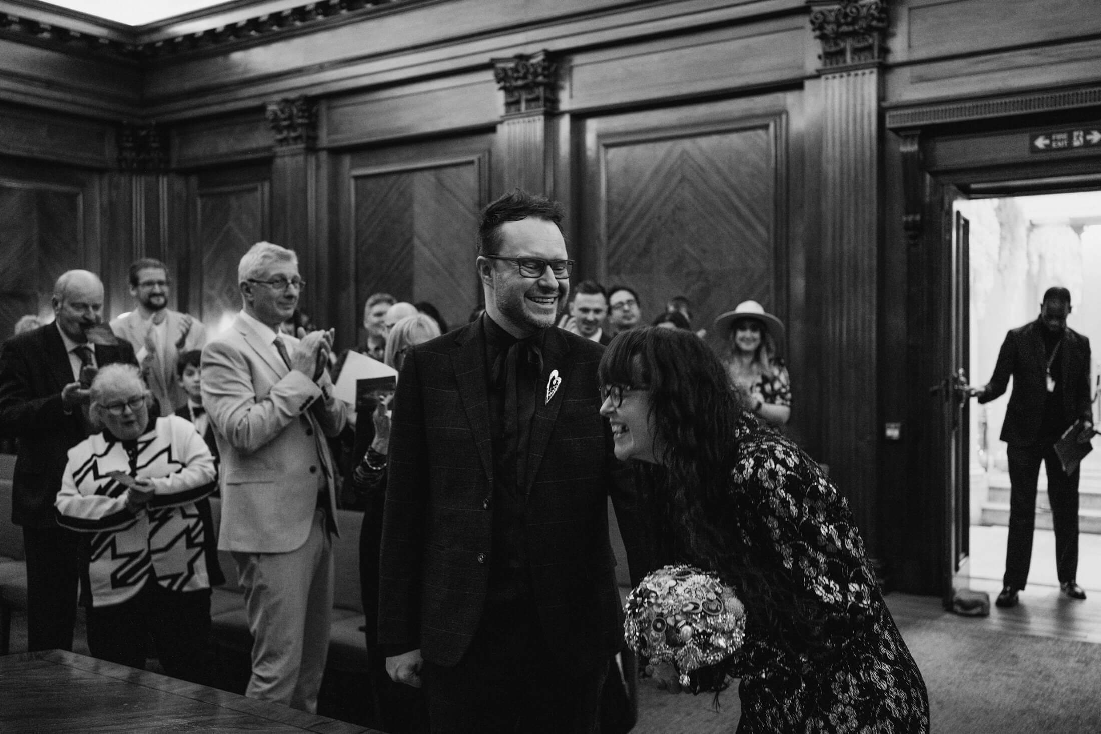 Bride and groom laughing as guests applaud inside wood-panelled hall during wedding ceremony.