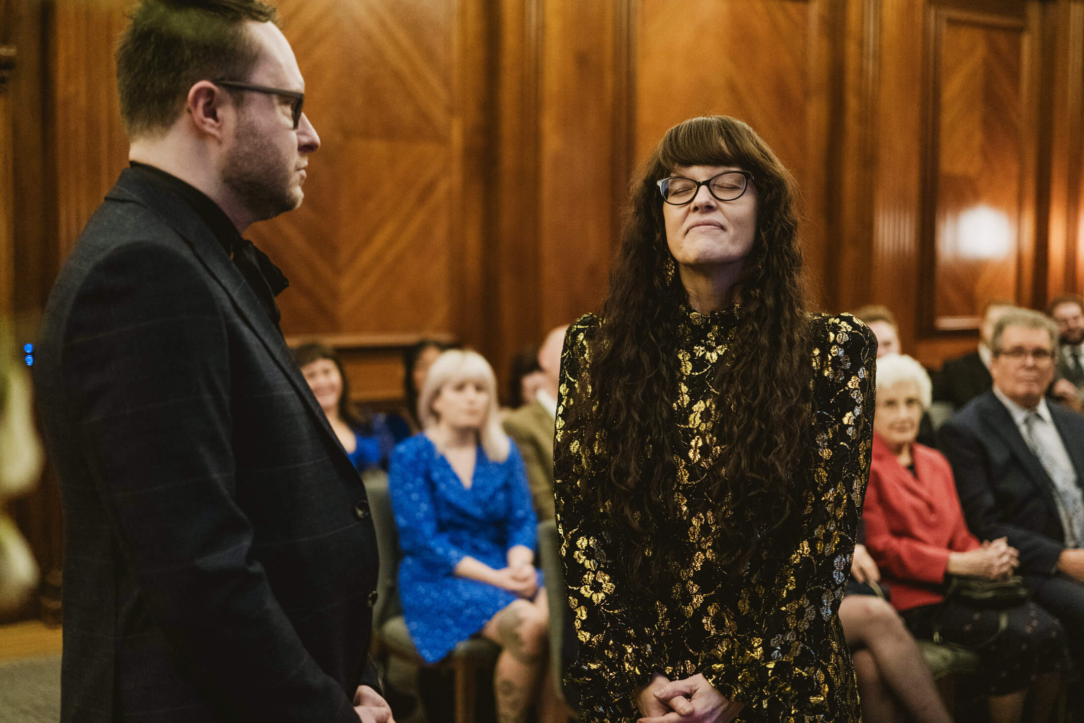 Woman closes eyes during wedding ceremony as partner stands beside her, guests watching softly.