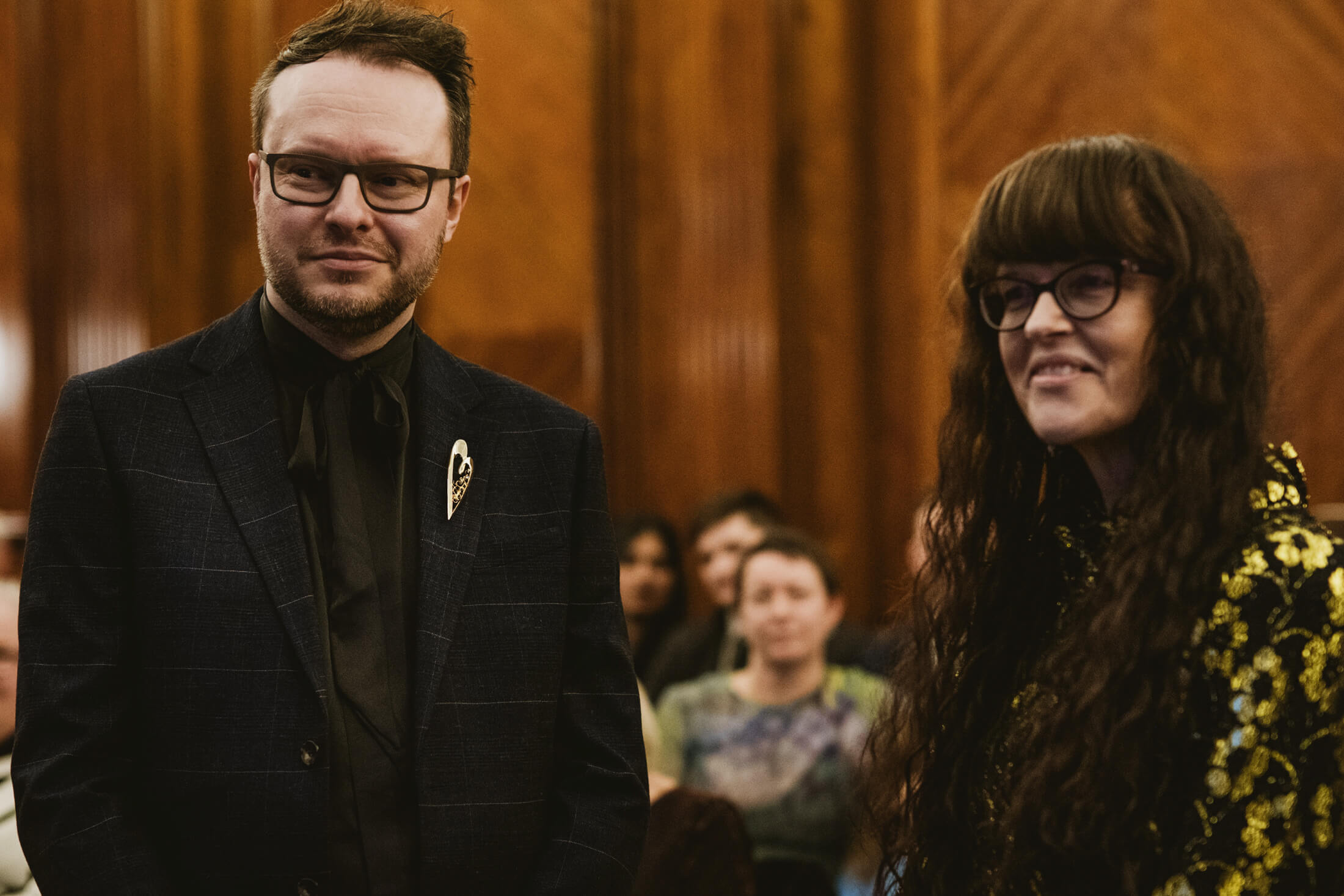 Couple stand together during wedding ceremony in wood-panelled hall, guests watching softly behind.