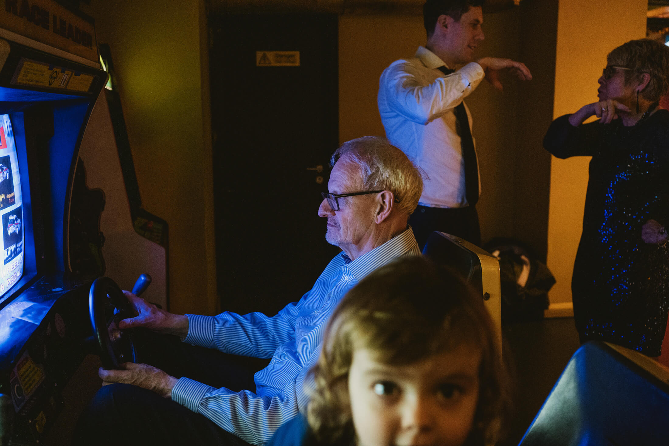In warm glow, older man plays arcade racing game as family chat nearby at wedding reception.