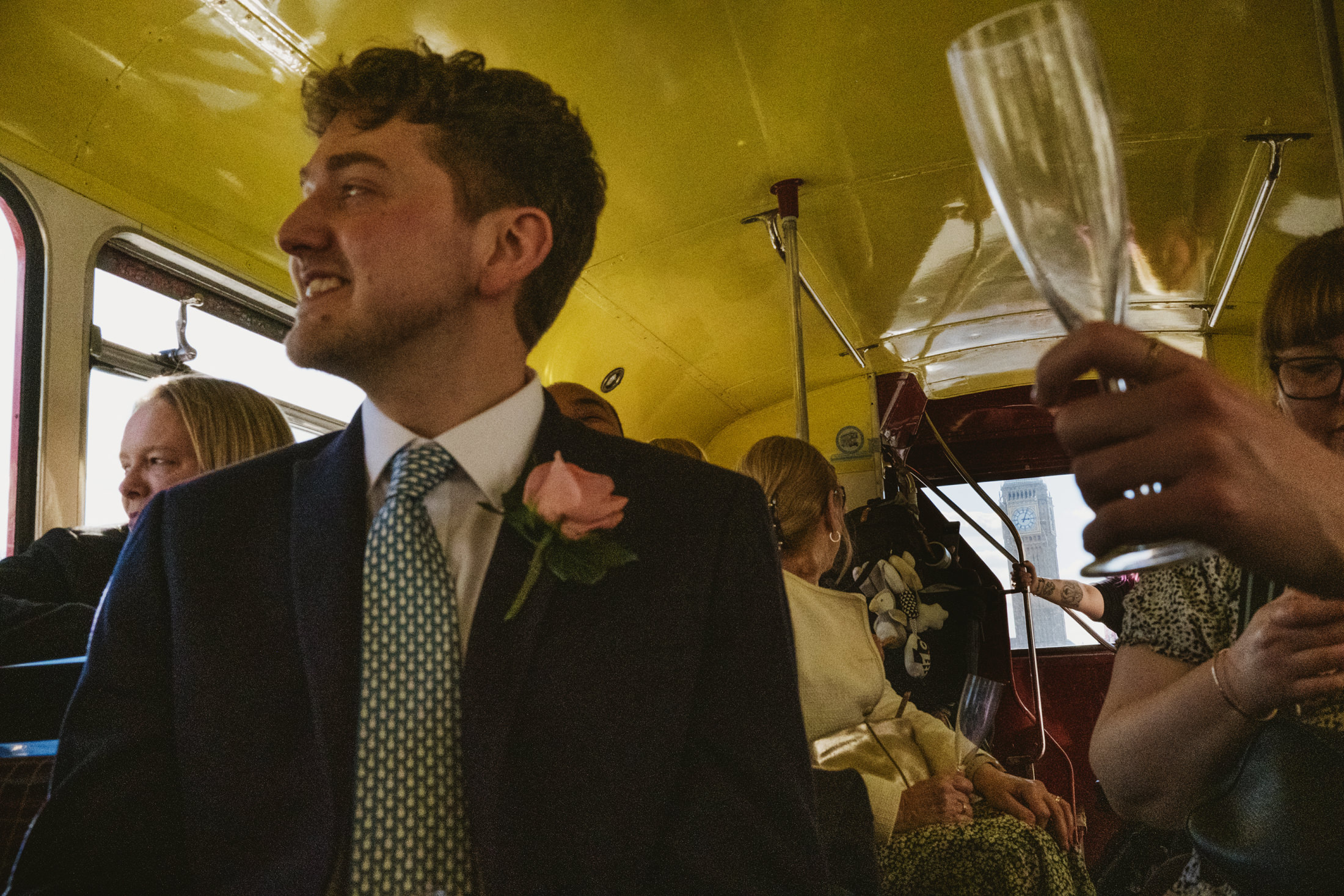 Groom smiling on vintage bus, guests holding champagne, Big Ben passing outside, documentary wedding photography.