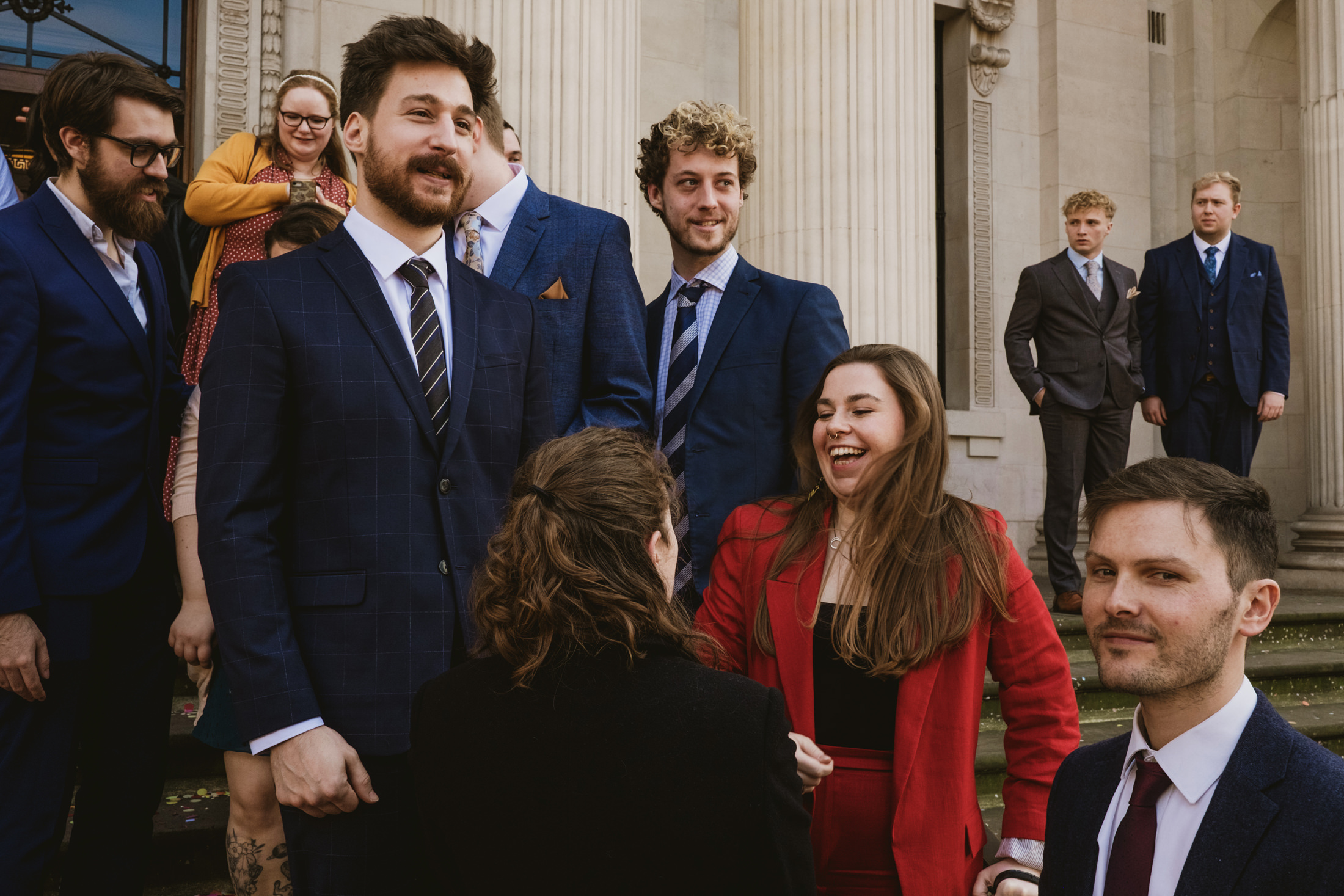 Wedding guests laughing on stone steps outside city hall, suits and bright red jacket catching afternoon light.