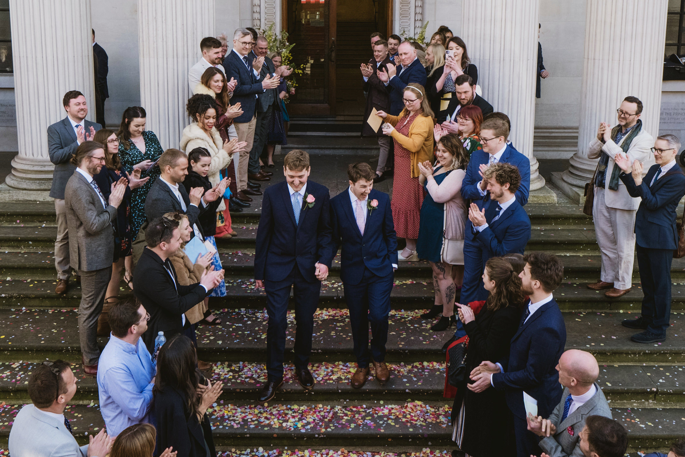 Couple walk through cheering guests as confetti falls on stone steps outside city wedding ceremony.