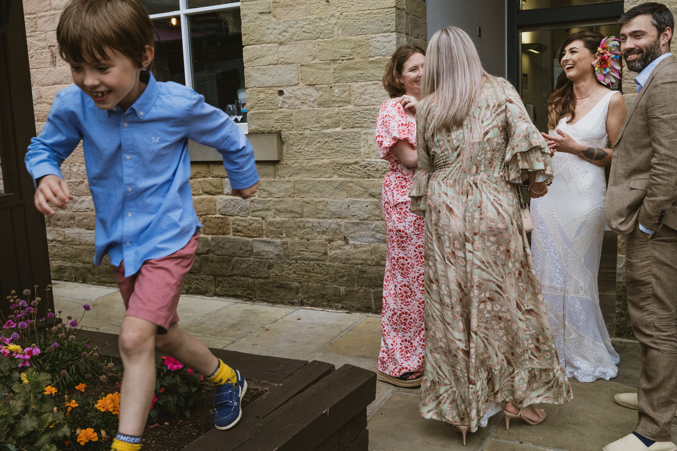 Boy runs past flowers as bride and guests laugh outside during documentary wedding photography.