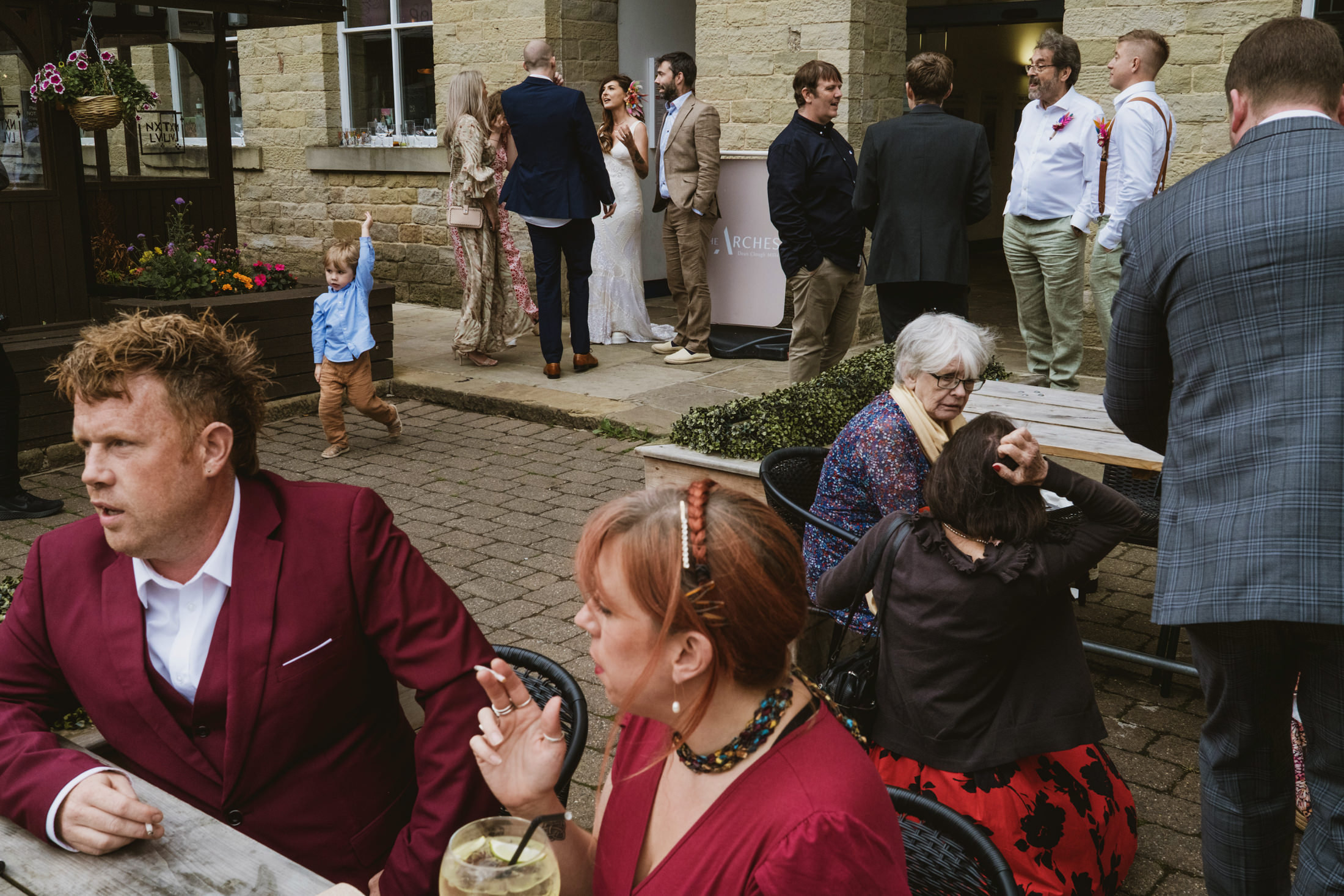 Guests chat outside stone venue as a child runs past, arm raised, during wedding photography.