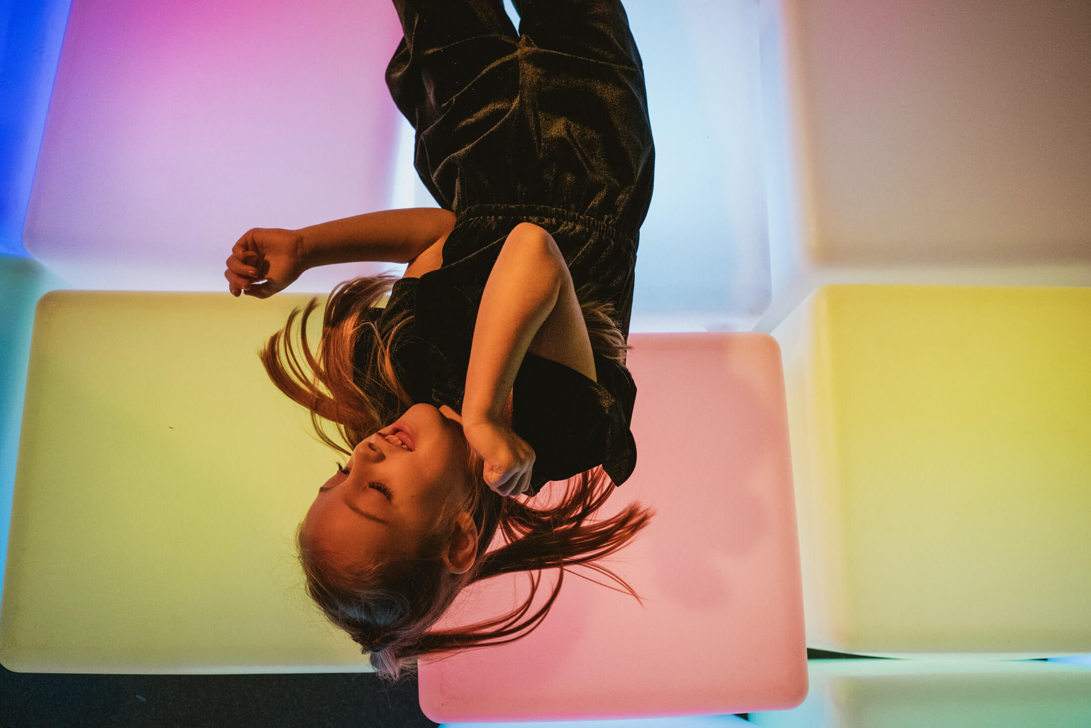 Child hangs upside down over pastel blocks, hair falling, smiling in soft coloured light.