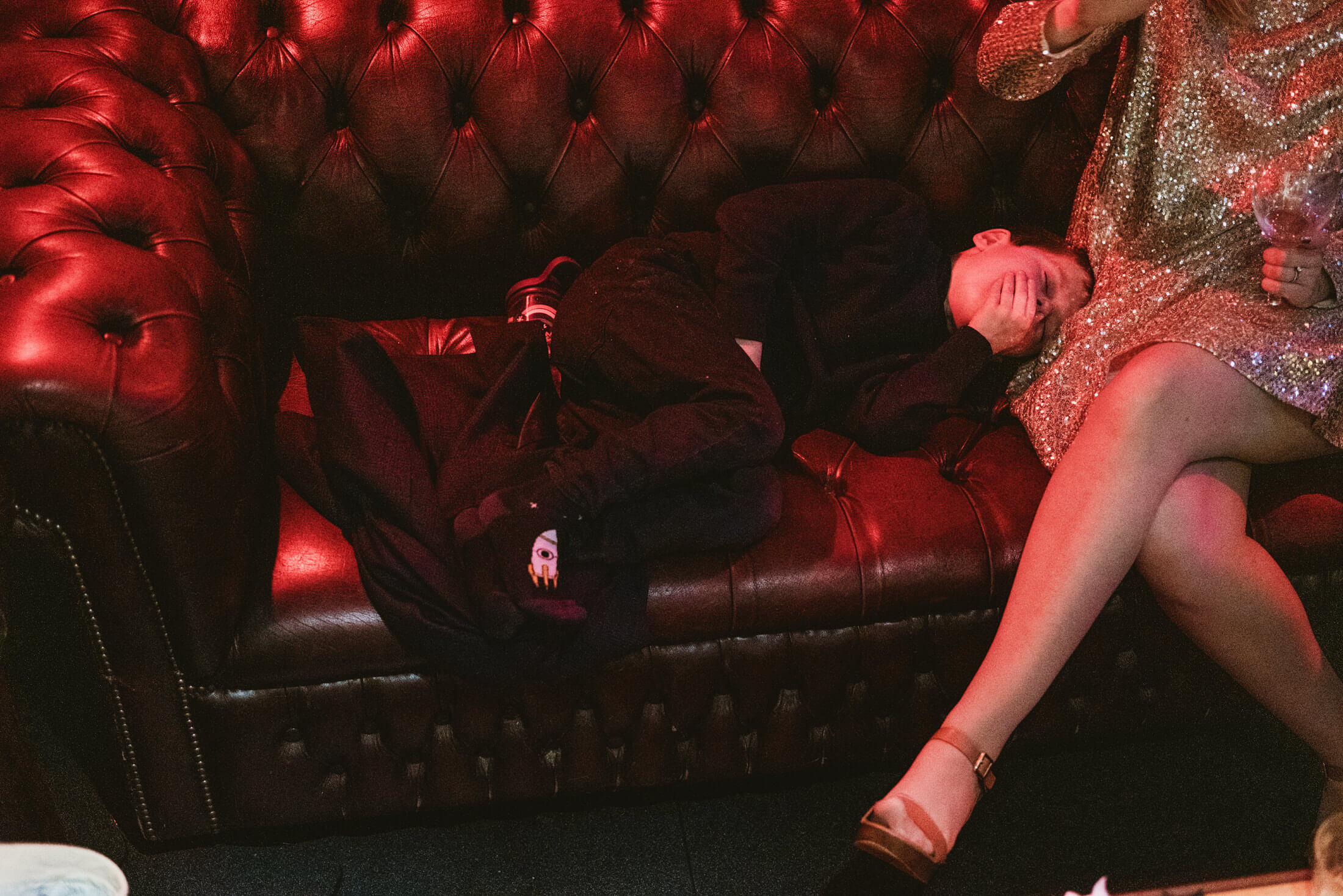 Child asleep on red leather sofa beside woman in glittering dress at wedding reception.
