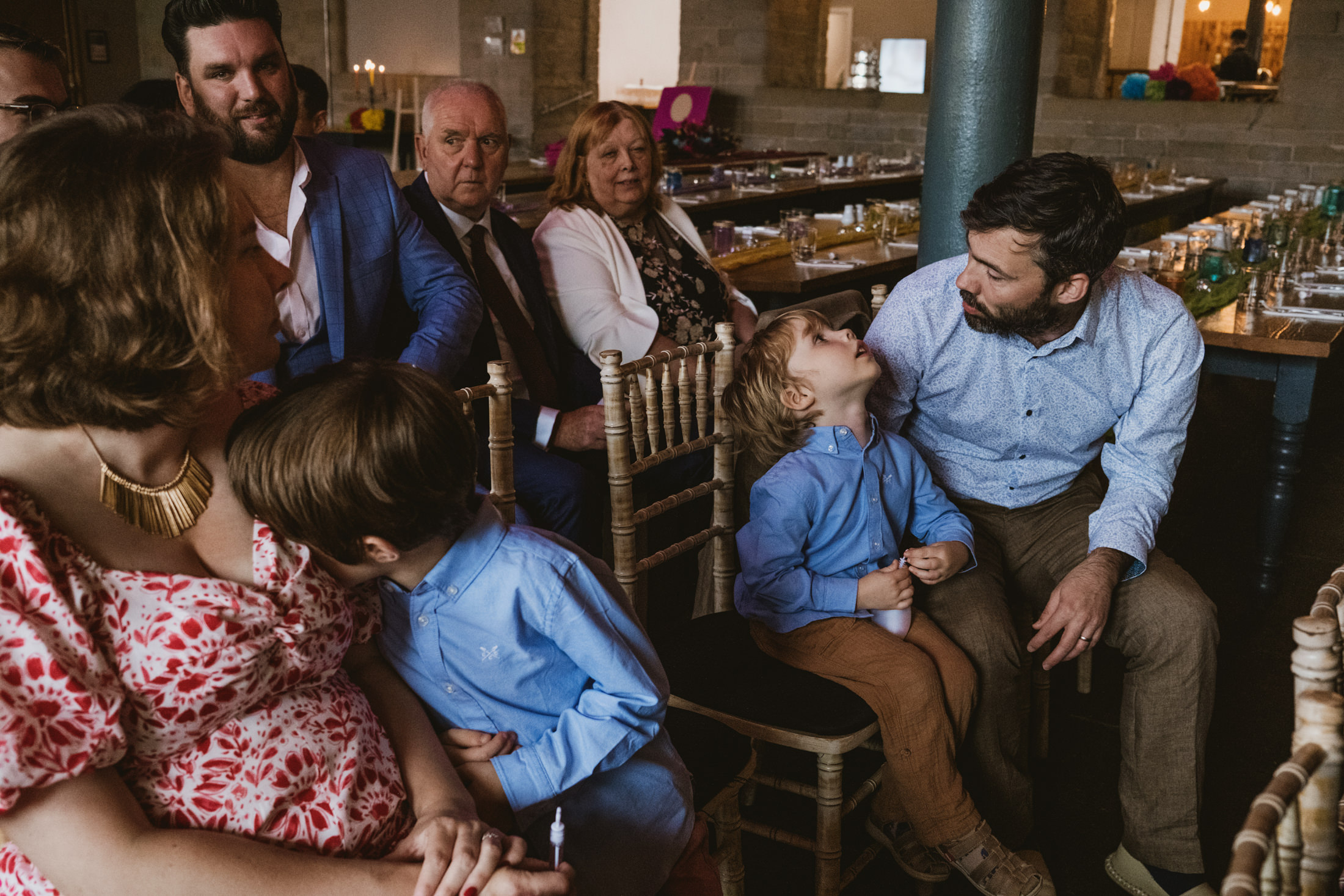 Father and young son share a quiet look among wedding guests at candlelit reception.