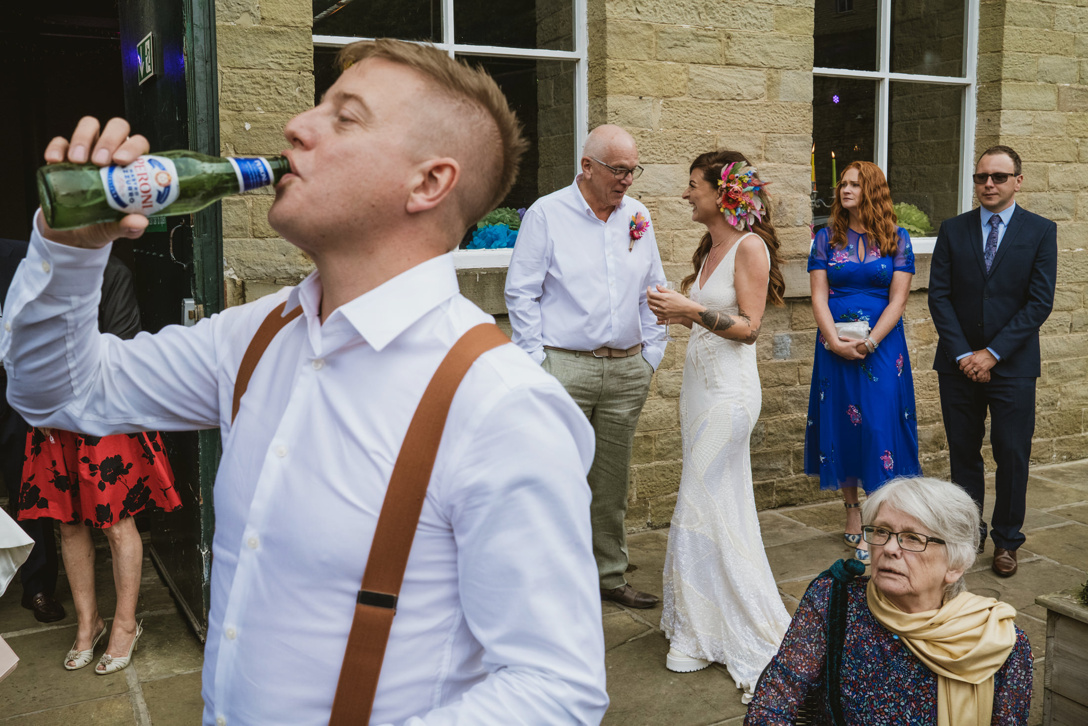 Guest drinks from bottle as bride laughs with family outside stone venue, documentary wedding photography.