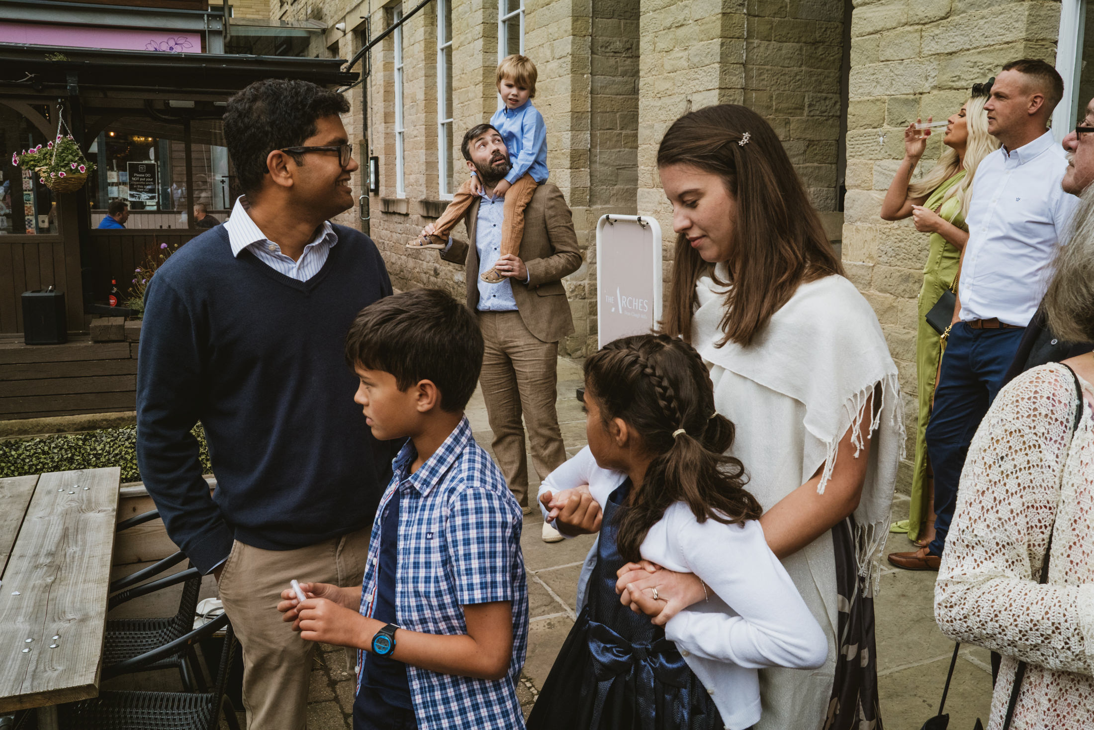 Guests chatting outside stone venue, father holding child aloft during relaxed documentary wedding photography moment.