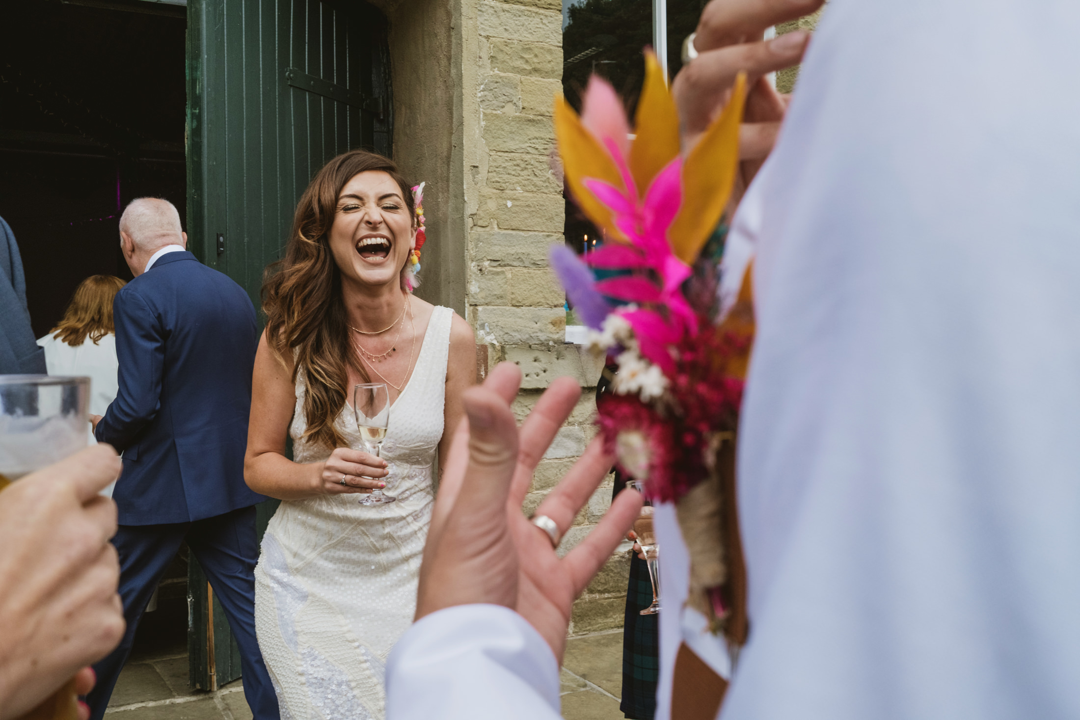 Bride laughing with champagne outside stone doorway, guests and bright bouquet blurred in foreground – documentary wedding photography.