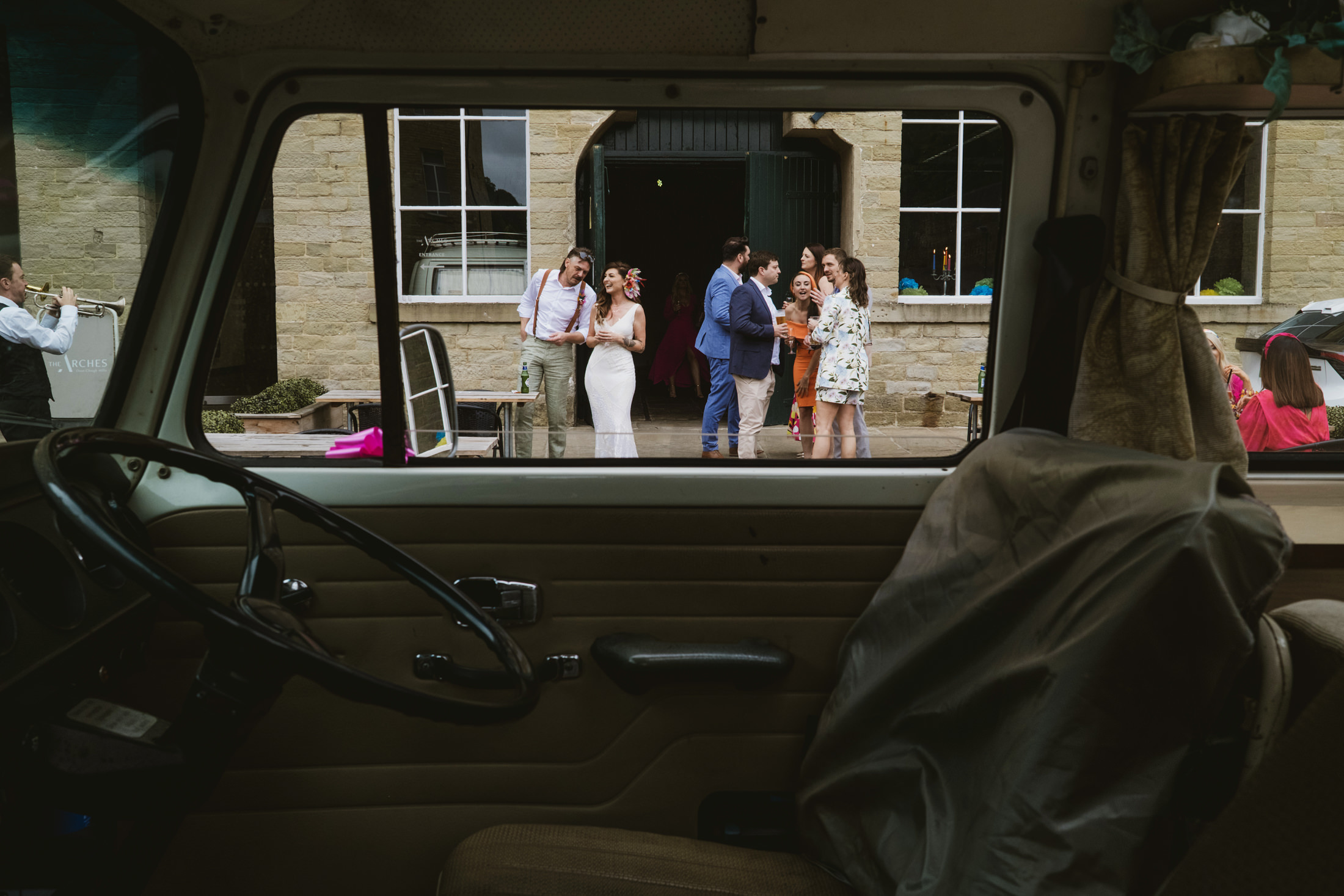 Guests laughing outside stone venue, seen through campervan window during documentary wedding photography.