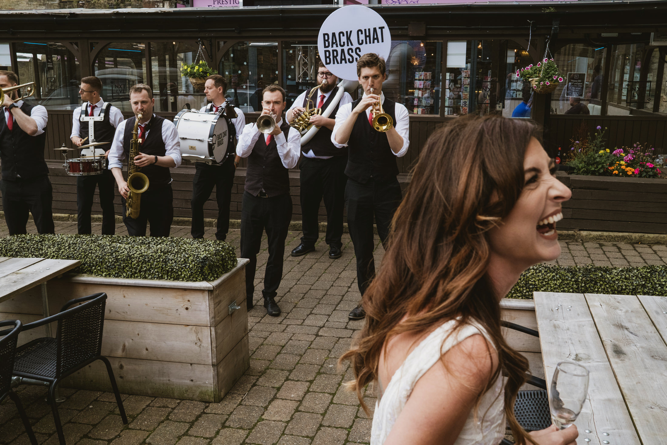 Bride laughing as brass band plays outside pub, champagne in hand.