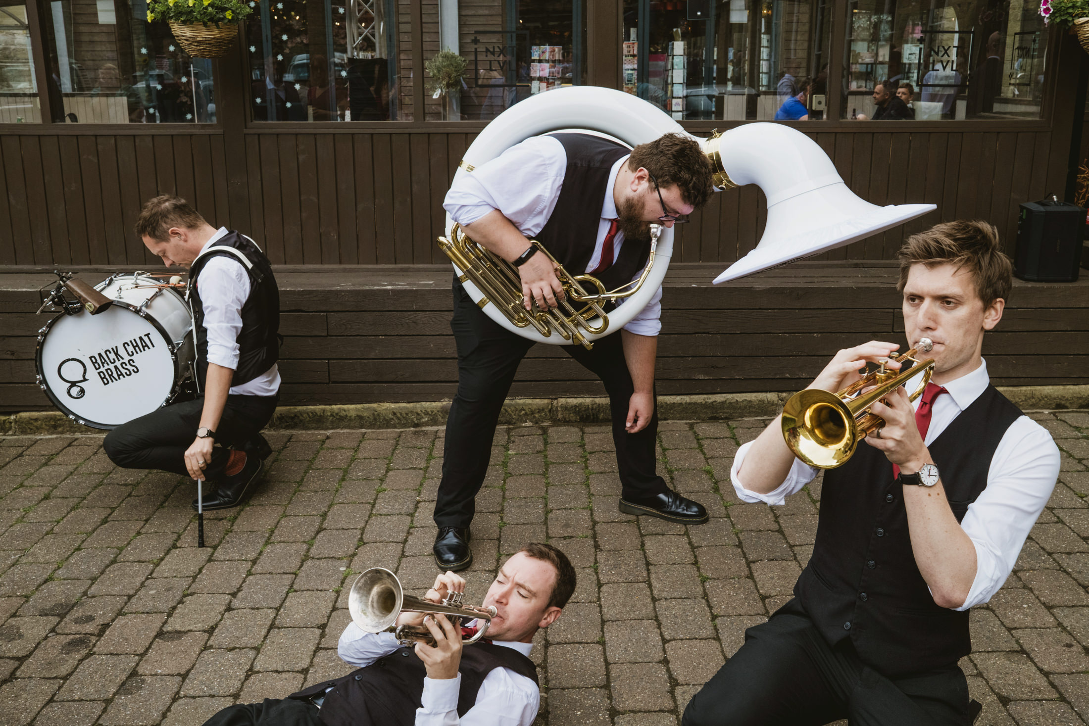 Brass wedding band playing on cobbled street, one musician lying down as others perform outside pub.