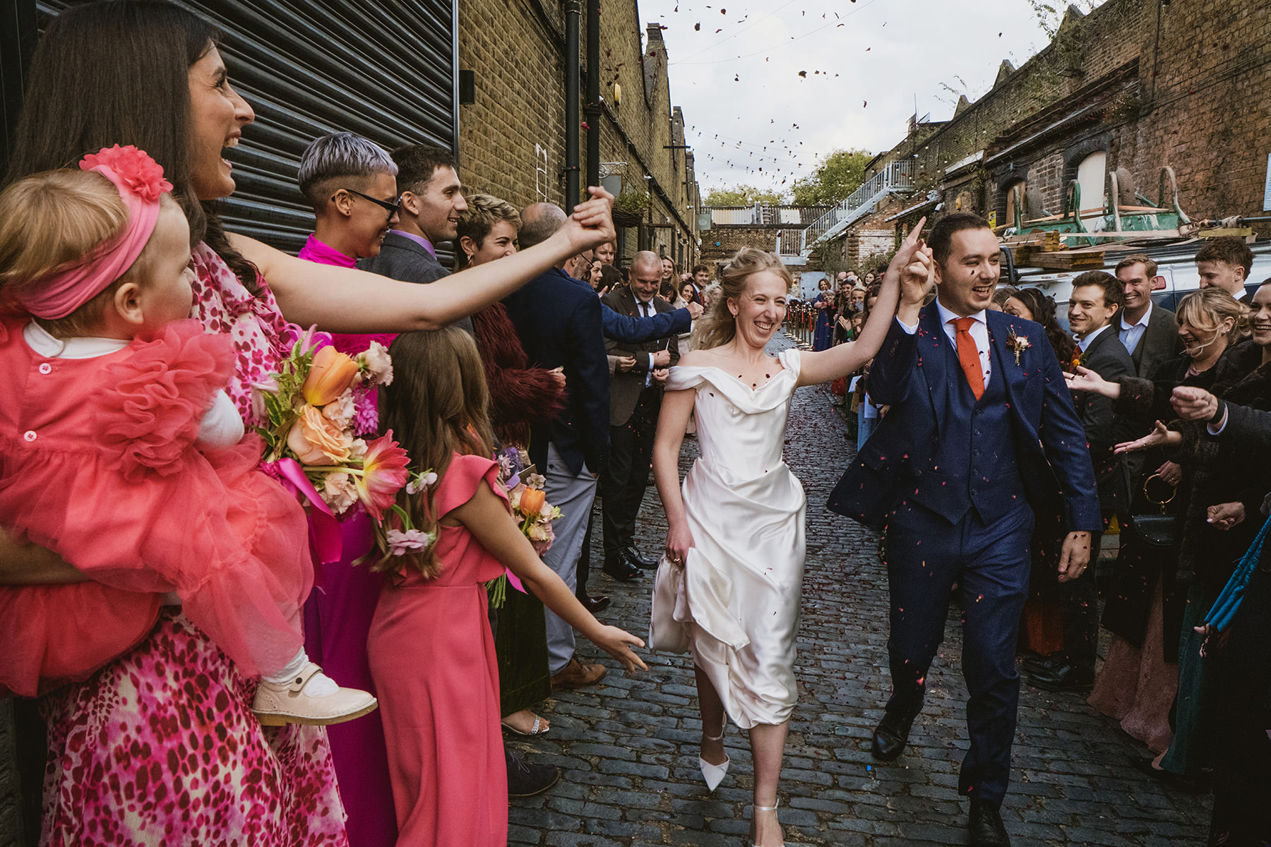 An energetic, unposed confetti shot of a wedding couple running through a cloud of petals in the historic tram depot alleyway at Clapton Country Club, East London.