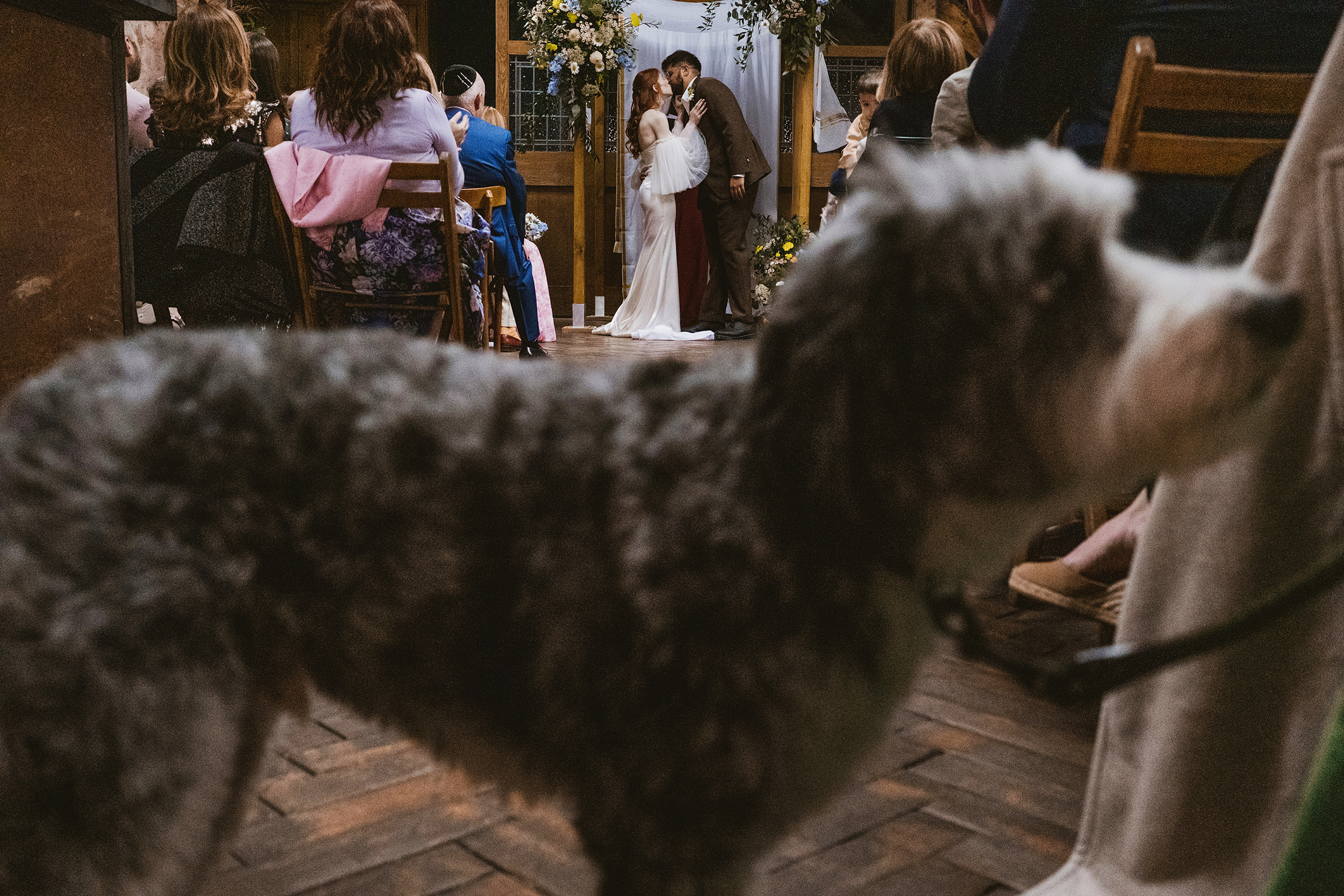 A documentary-style wedding photograph at Clapton Country Club capturing the couple's first kiss, framed by their dog in the foreground against the industrial brickwork of the Department of Life