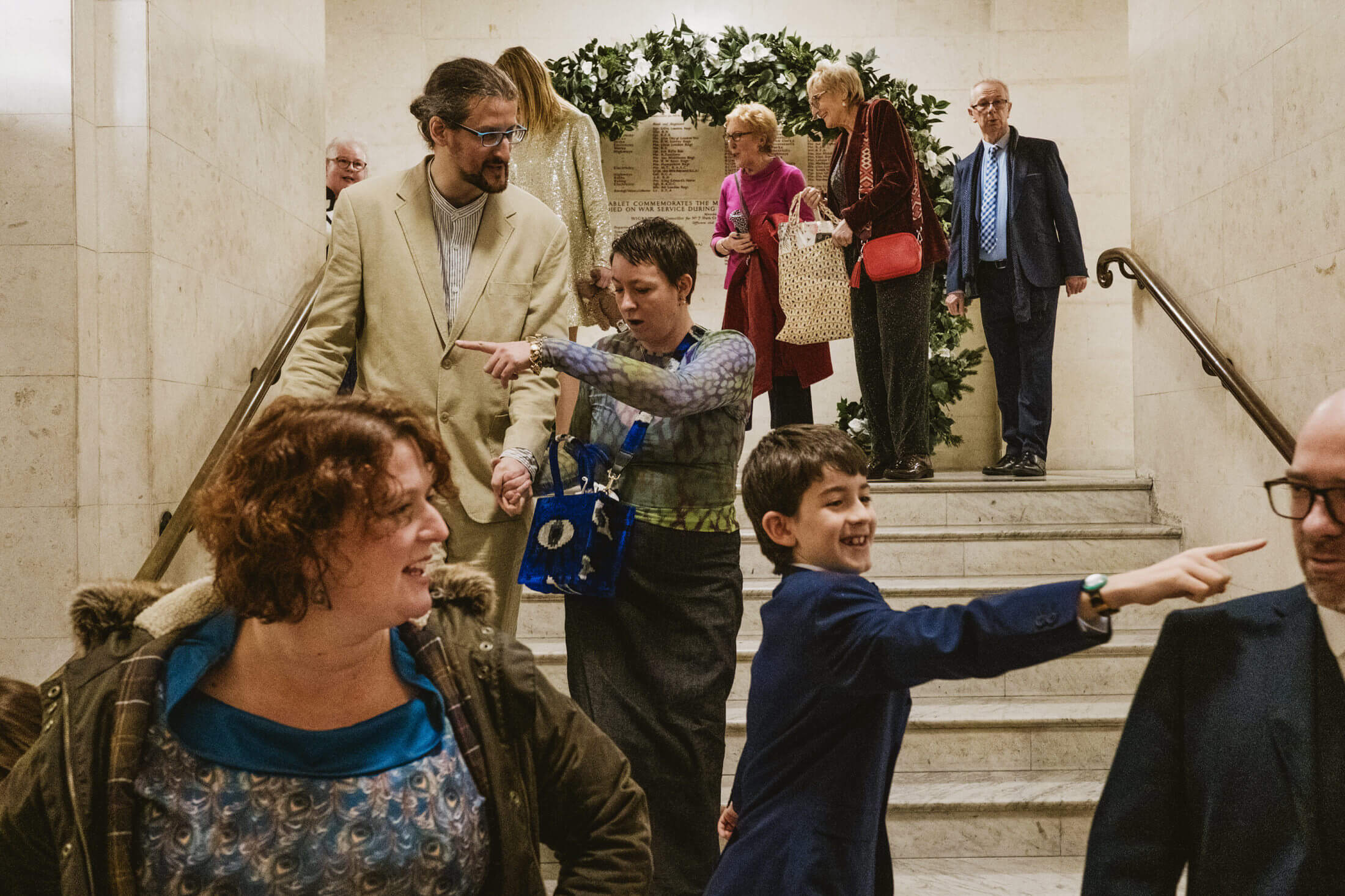 Wedding guests laugh and point while gathering on marble steps beneath a leafy arch.