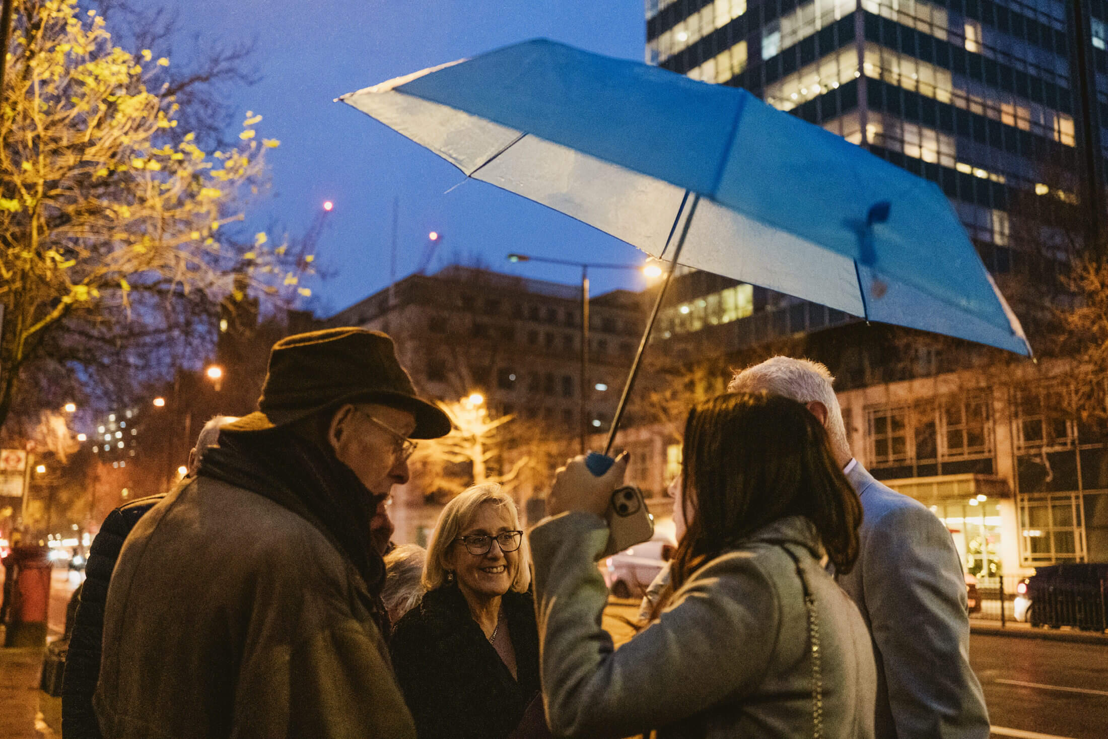 Guests gather beneath a blue umbrella, laughing on a city street at dusk, documentary wedding photography.