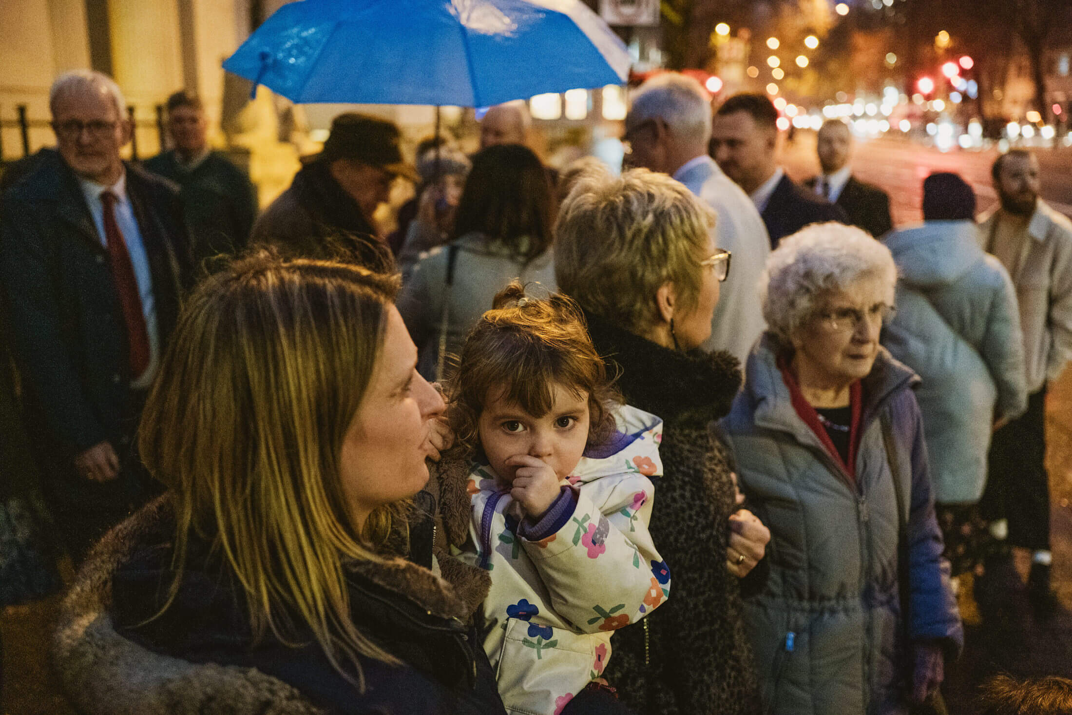 Woman holds child beneath blue umbrella as guests gather on rain-lit street at dusk, documentary wedding photography.