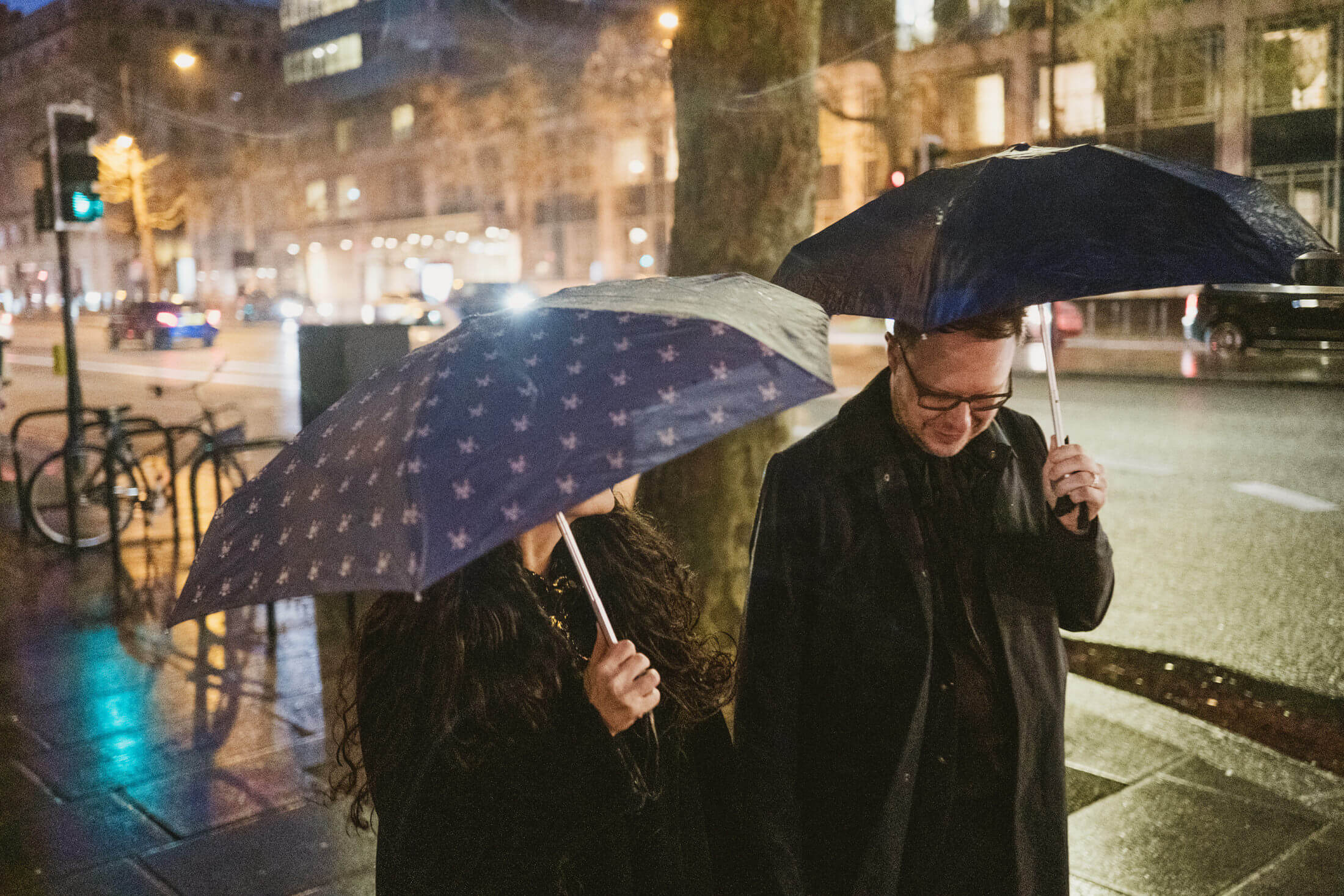 Couple walking beneath umbrellas on rain-soaked city street at night, sharing a quiet smile – documentary wedding photography.