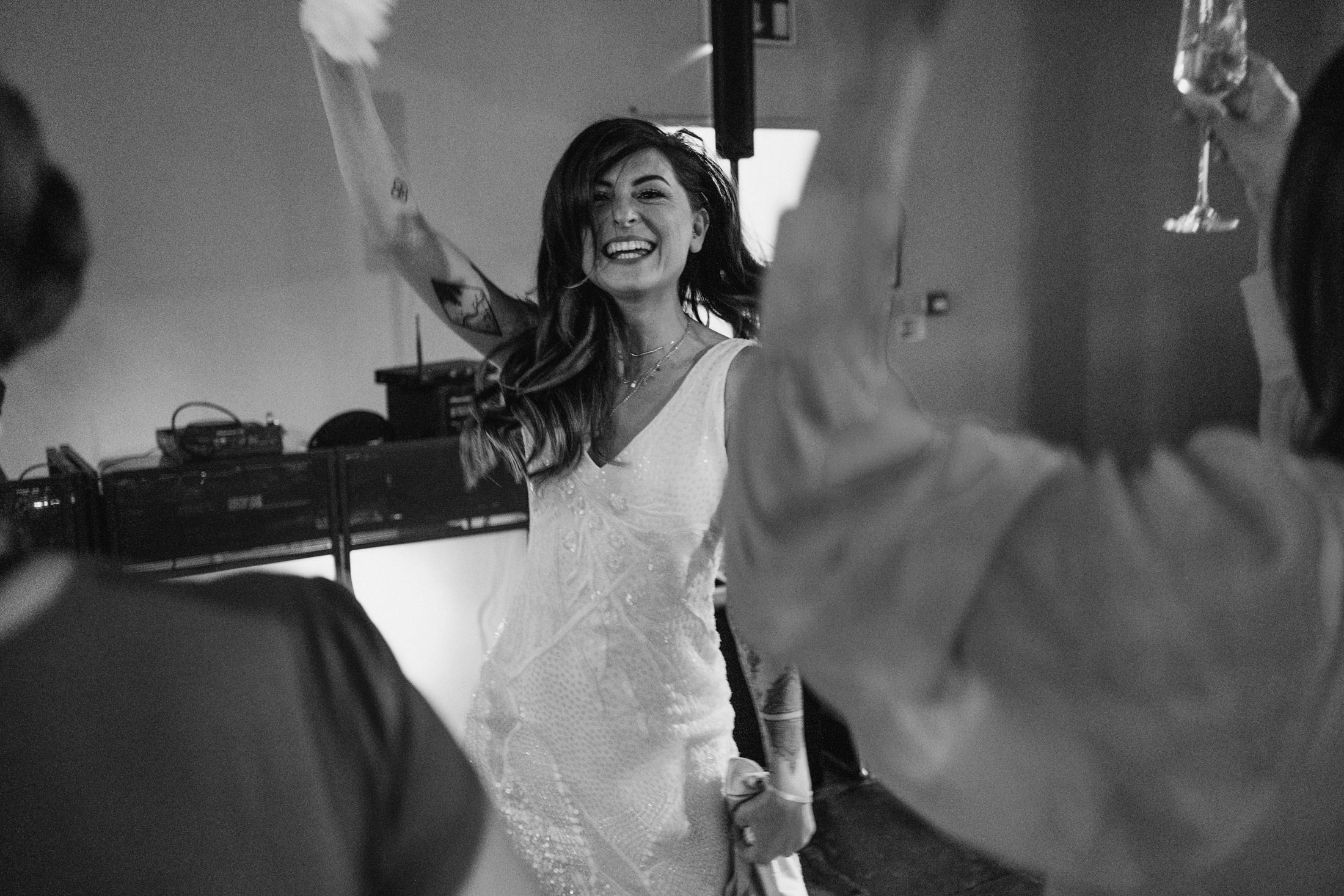 Bride laughing with arms raised on dance floor, guests blurred around her, documentary wedding photography moment.
