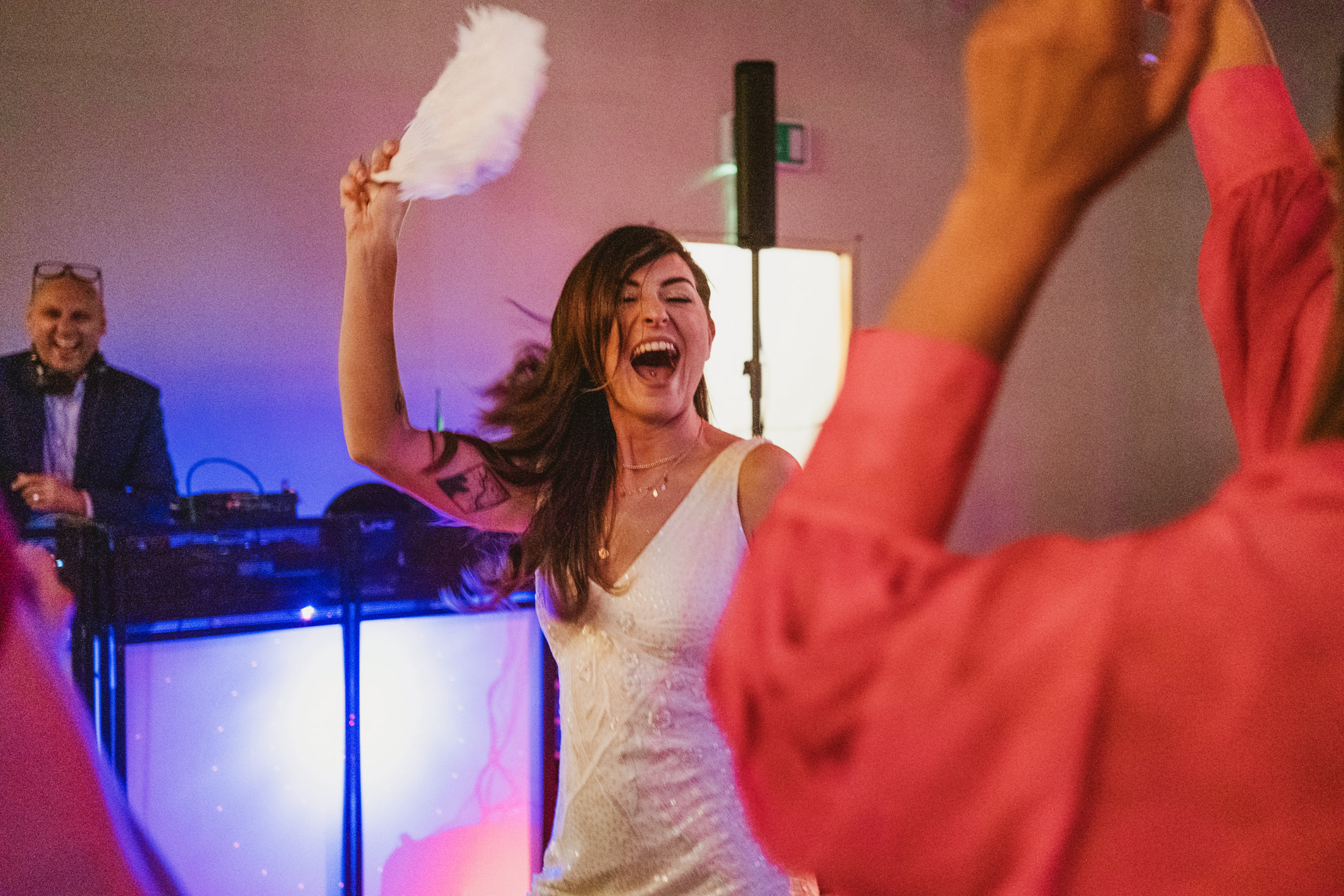 Bride laughing on dancefloor, waving feather fan as guests cheer during wedding reception.