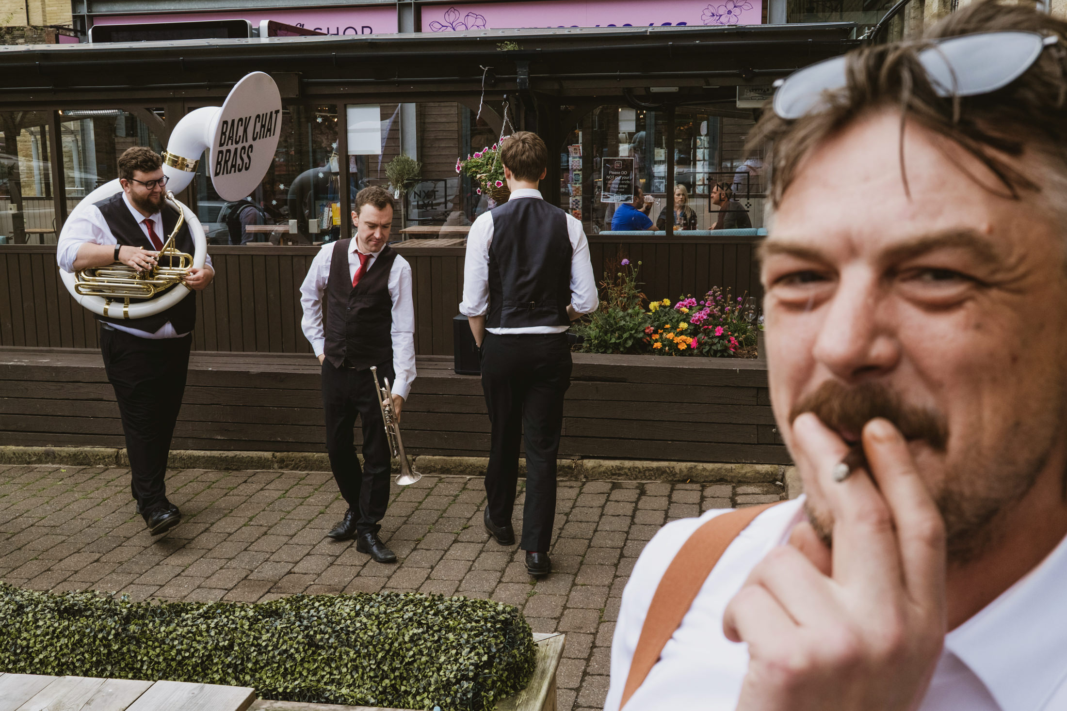 Brass band members gather outside a pub, one man smoking in foreground, during a lively wedding celebration.