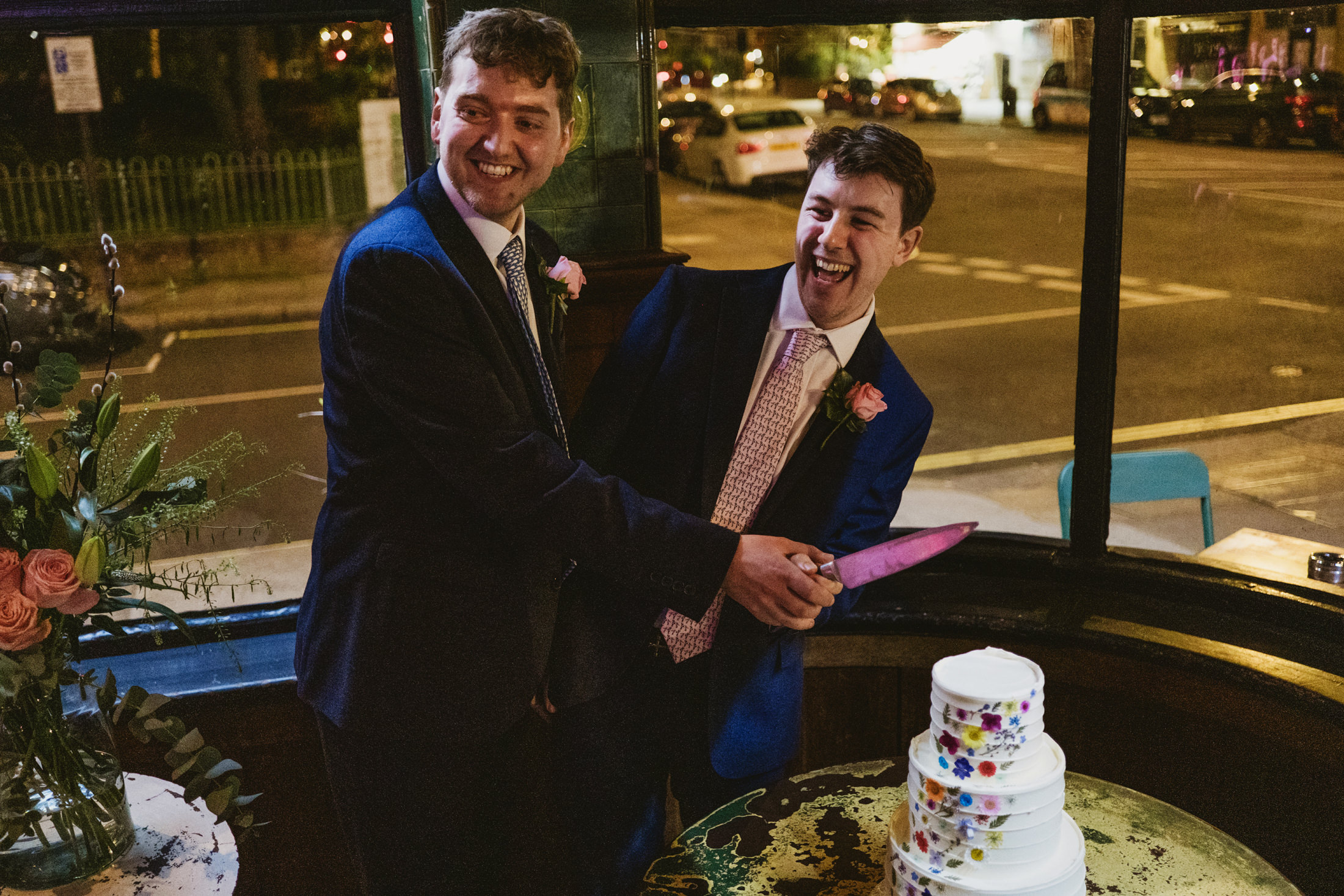 Two men in suits laugh while cutting a floral wedding cake by lamplight.