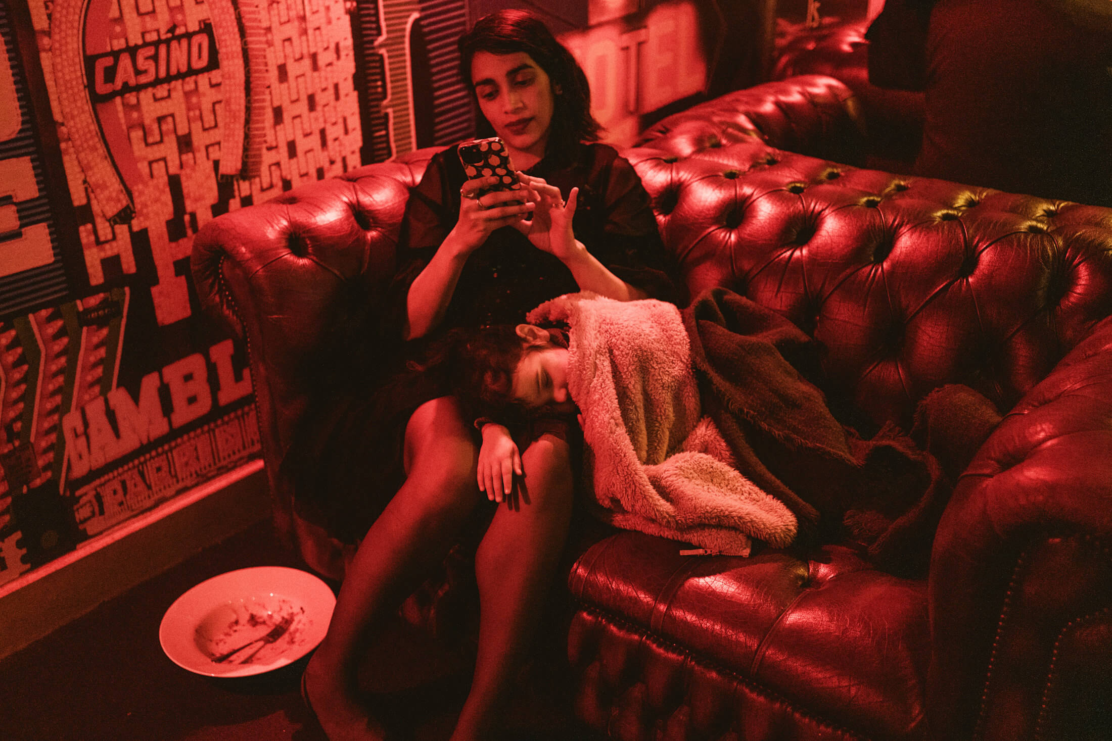 Woman checks her phone as a child sleeps beside her on a red leather sofa.
