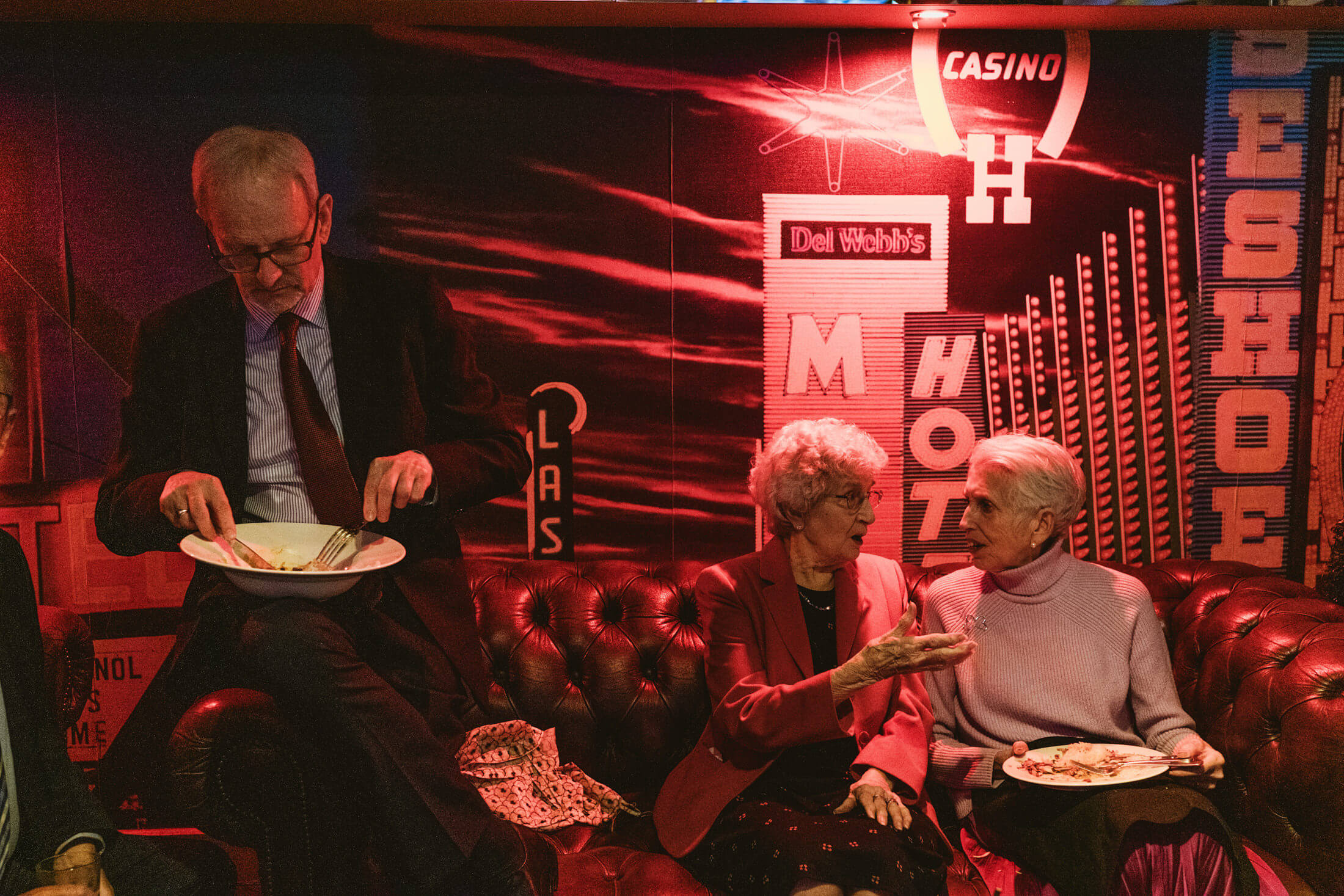 Elderly guests share food and conversation on a red leather sofa beneath glowing neon signs at a wedding reception.