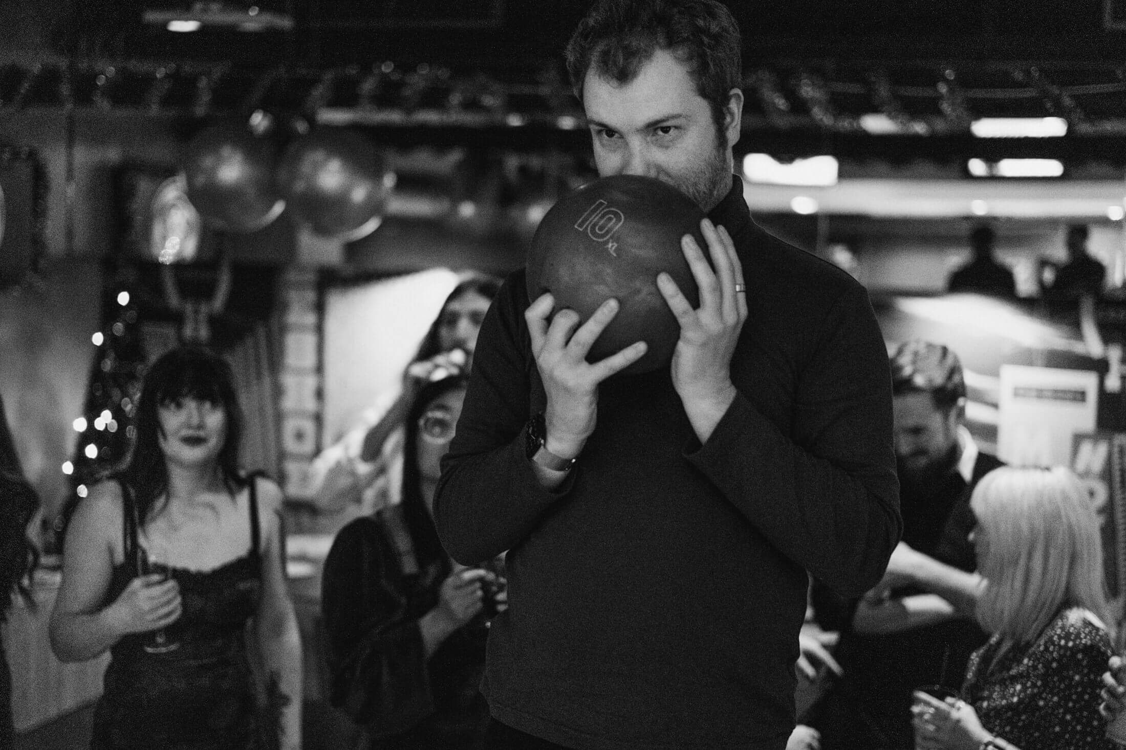Man lifts bowling ball to his face as friends watch at lively wedding reception.