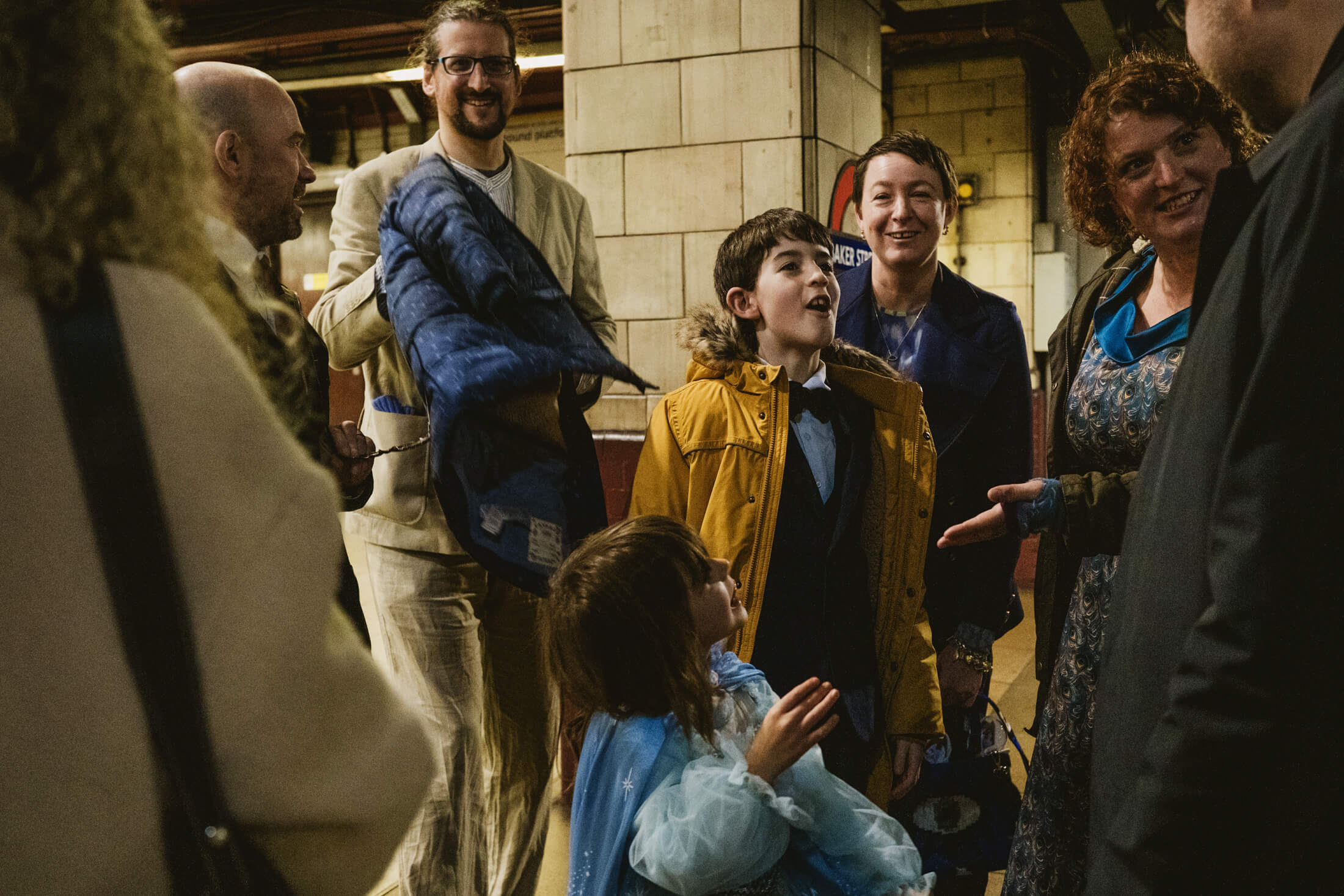 Guests laugh together on an underground platform, children in bright coats chatting during a wedding celebration.
