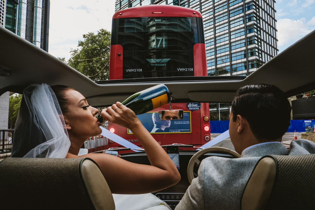 Bride drinks champagne in car behind London bus.