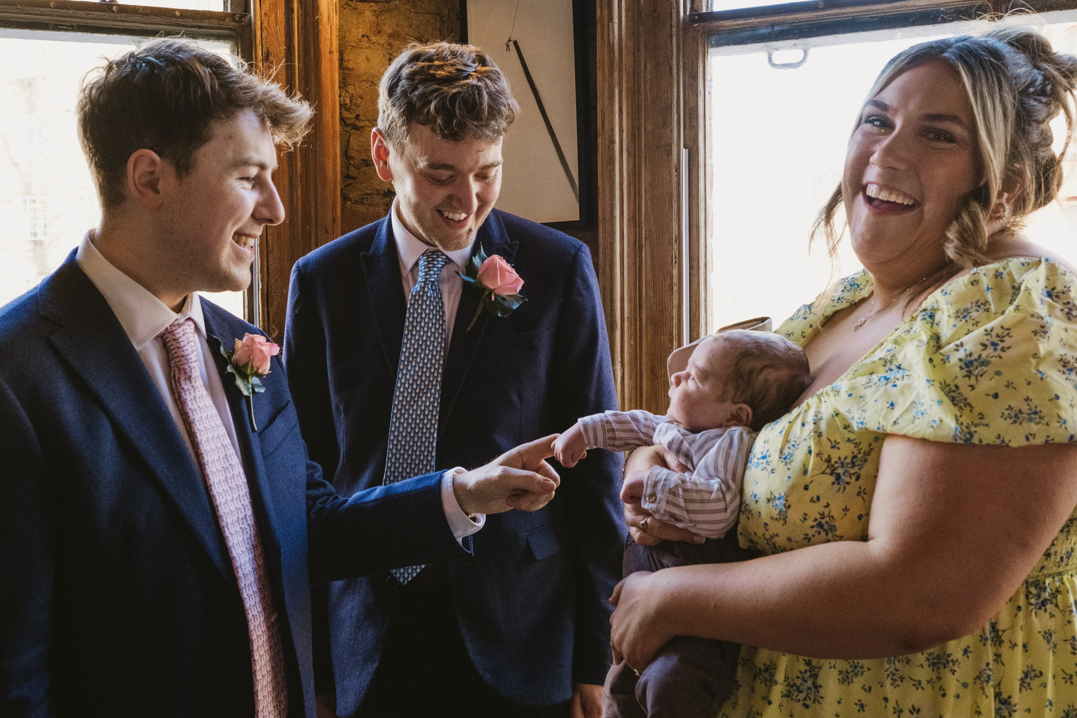 Two suited men smile at baby in woman's arms by window, soft laughter during documentary wedding photography.