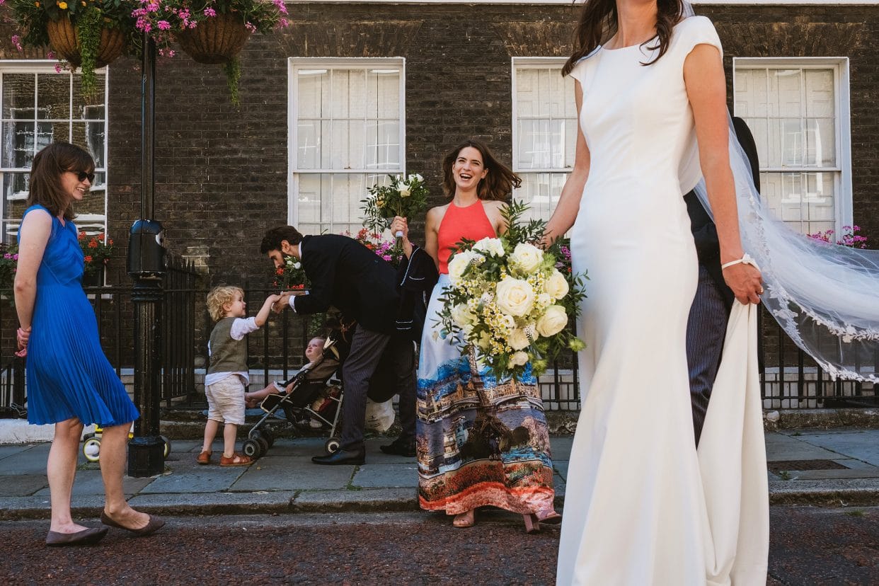 layered micro wedding street scene in London. In the foreground we can see the bride in her dress, holding her train in one hand and bouquet in the other. Behind and left is a bridesmaid in a multi-coloured dress holding a bridesmaid bouquet. Left of her and furthest away the groom bends down and holds the hand of a young wedding guest with a child in a pram framed between them. On the far left of the image and on the same plane as the bridesmaid a girl in a blue dress and sunglasses stands with her arms behind her back