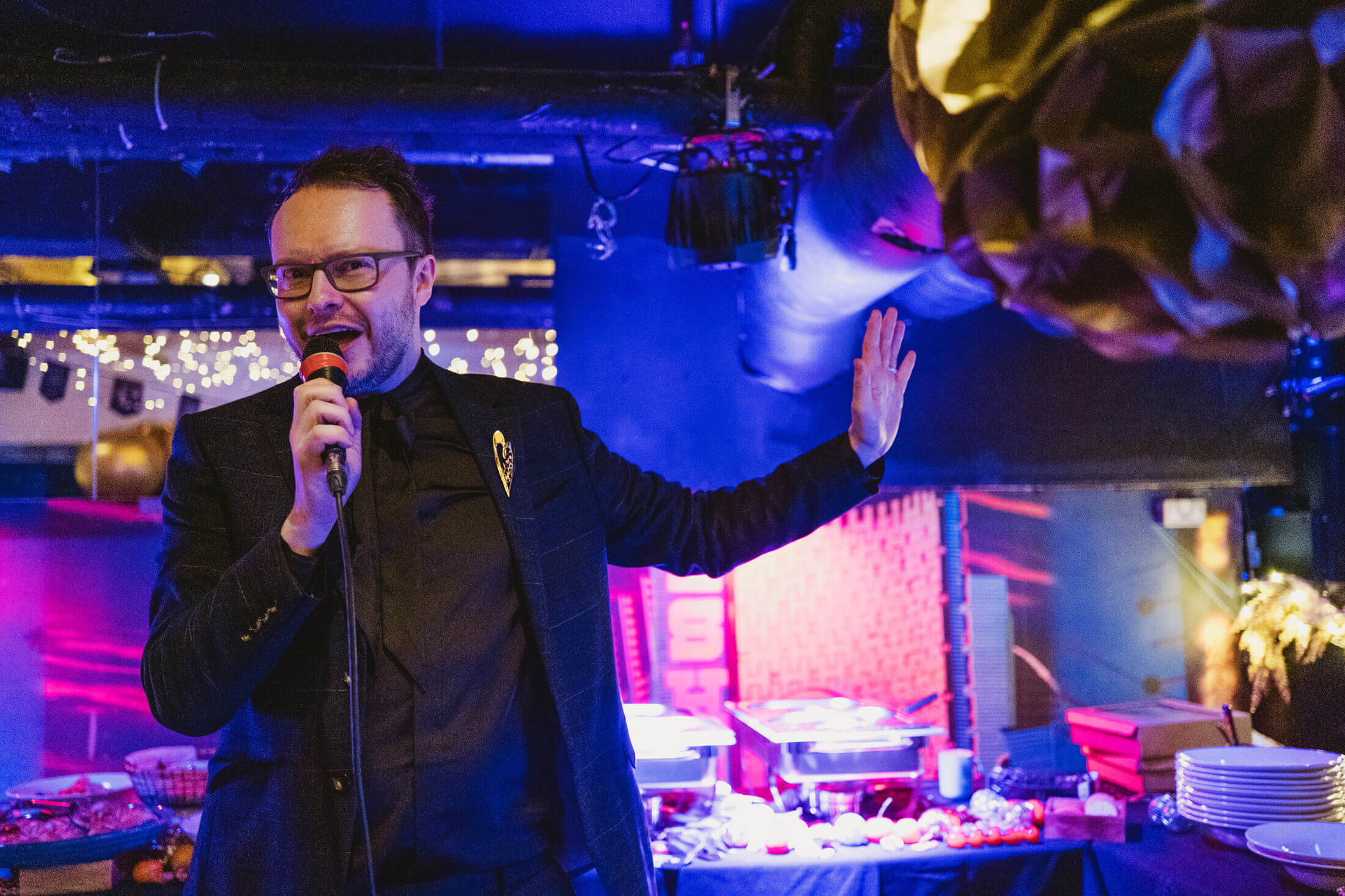 Man speaking into microphone at wedding reception, colourful lights glowing behind him.