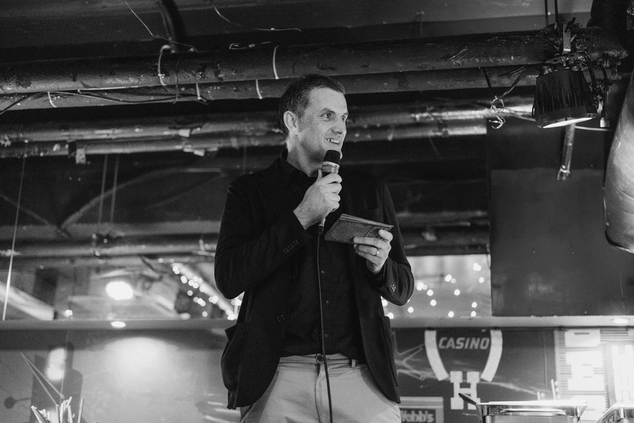 Man smiling while giving a wedding speech, holding notes beneath soft reception lights.