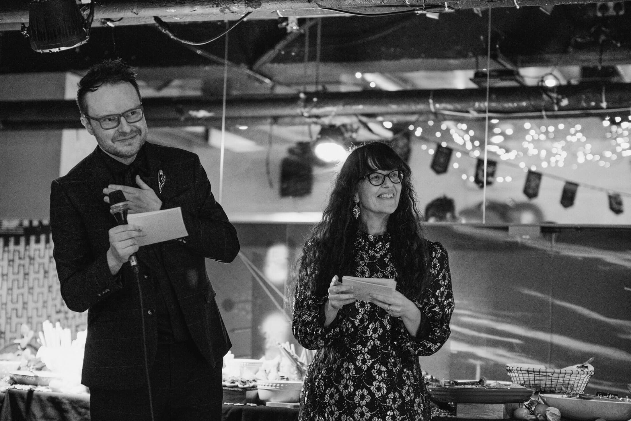 Two guests smile while giving a wedding speech beneath soft fairy lights, notes and microphone in hand.