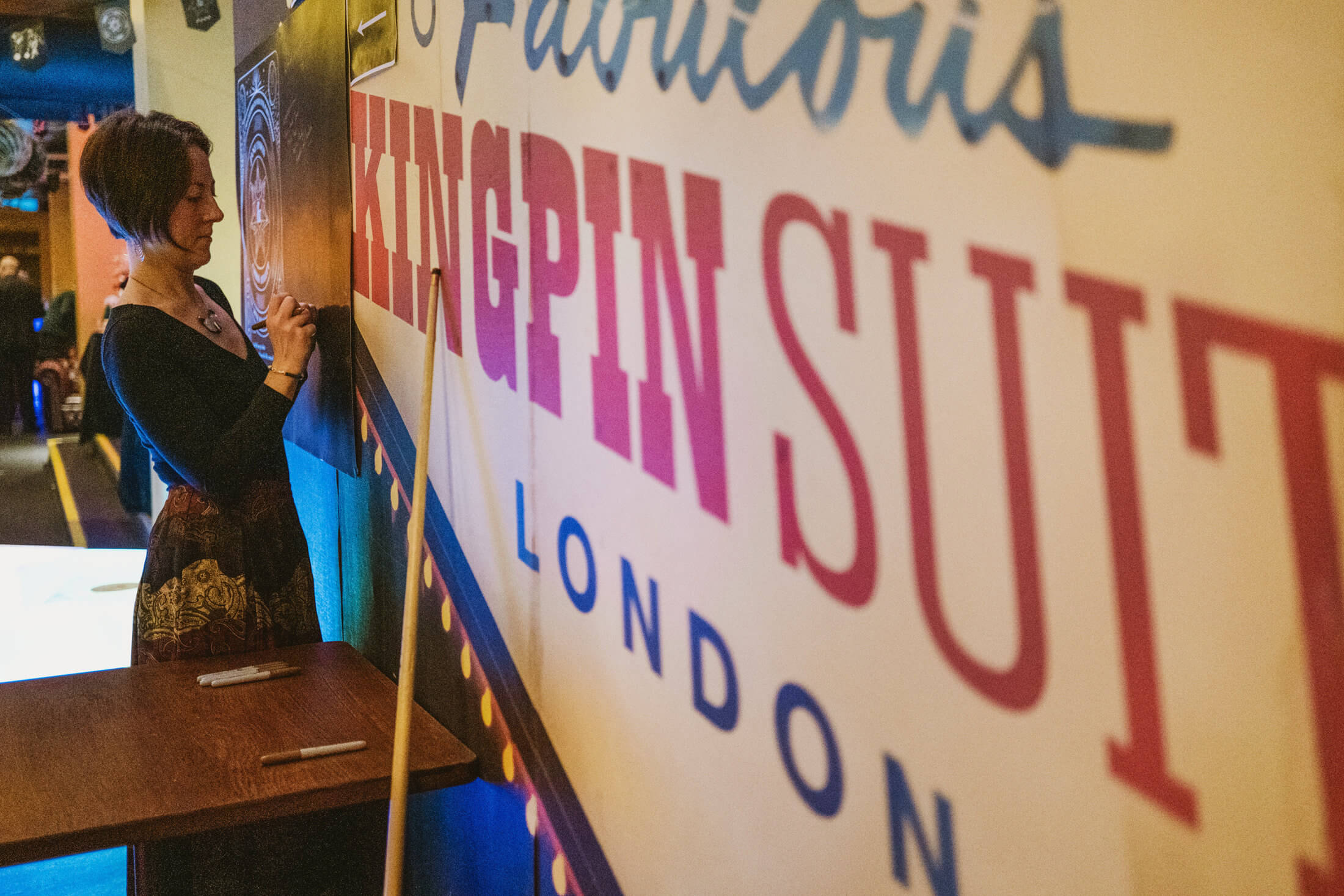 Woman writes on a large wall sign in a softly lit London venue, quiet concentration in profile.