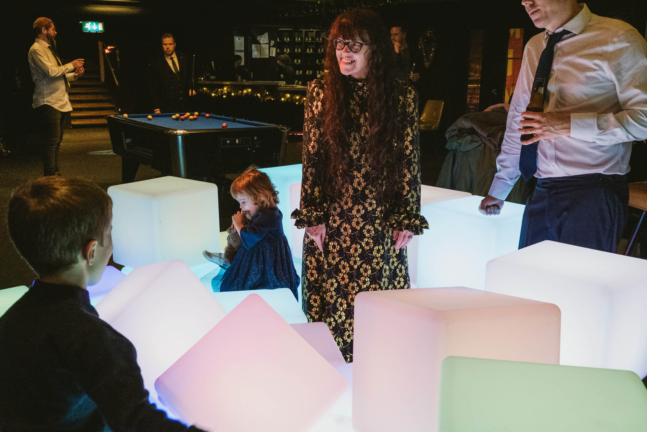 Guests laugh around glowing cubes as a child cuddles a toy during wedding reception.